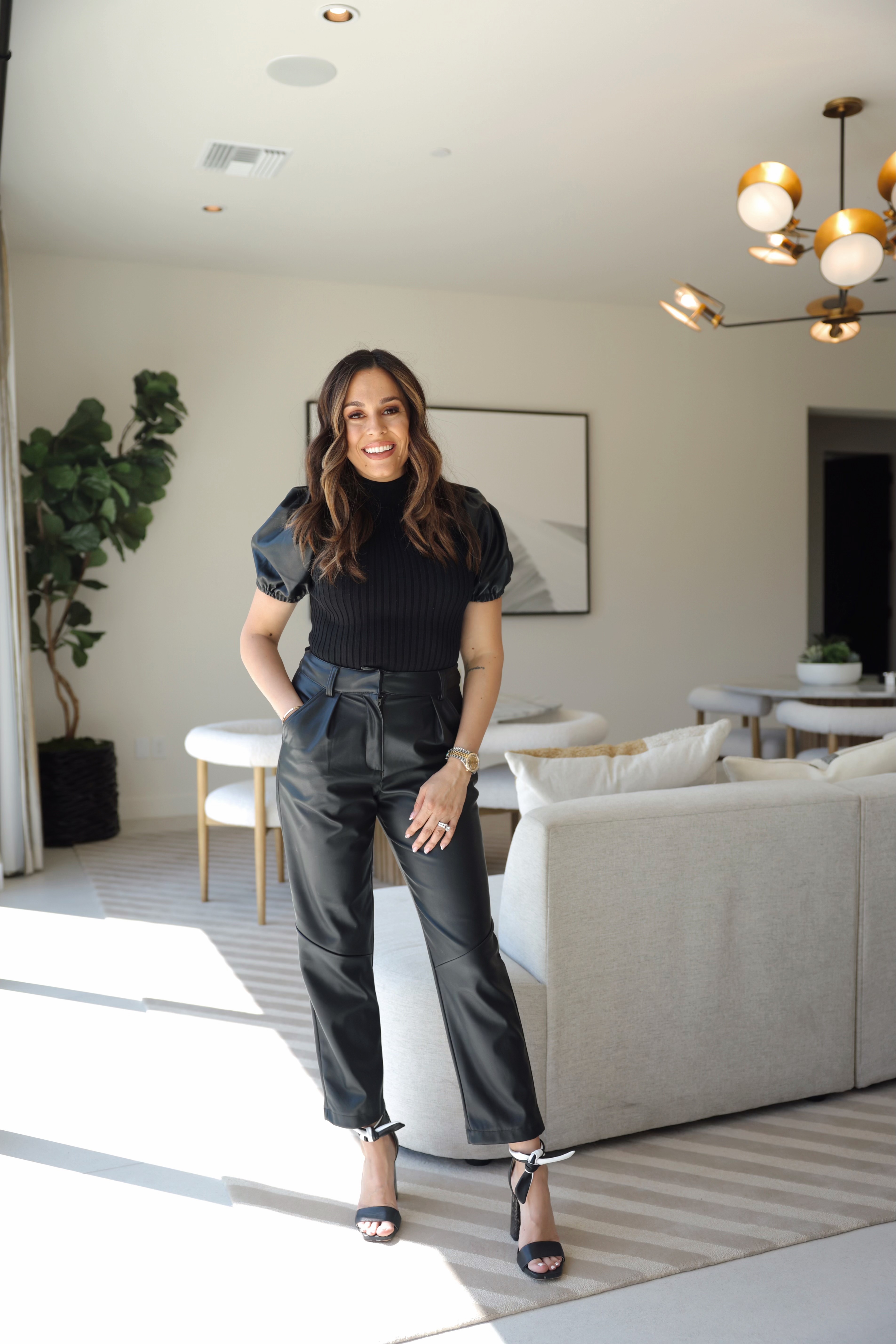 Woman with long hair in black outfit standing in a bright living room with a sofa, plants, and modern chandelier.
