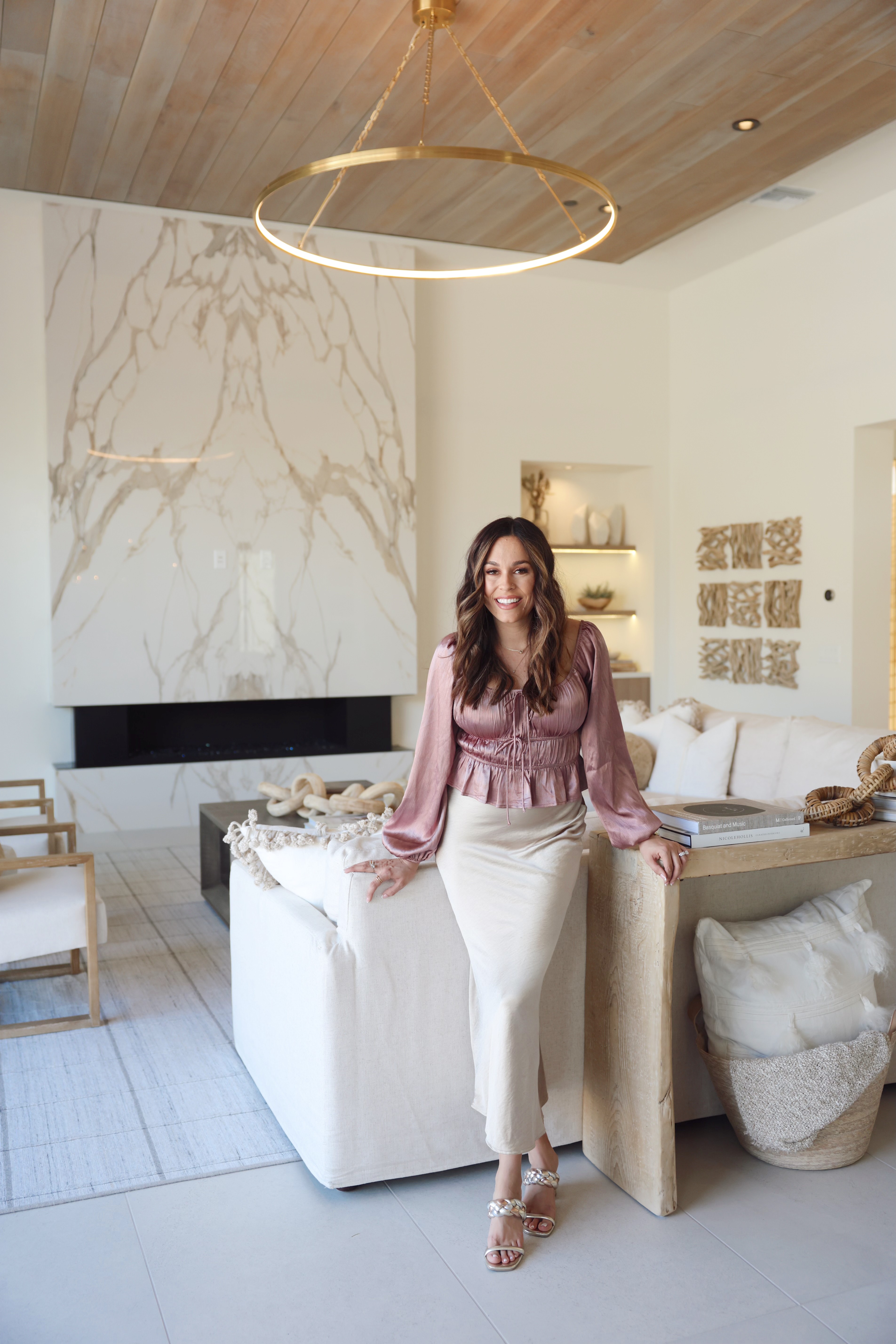 Woman in pink blouse and white pants standing in modern living room with marble fireplace and wooden ceiling