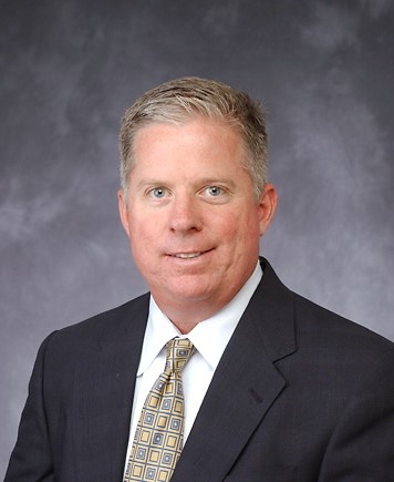 Portrait of a middle-aged man with short, light-colored hair, wearing a dark suit, white shirt, and patterned tie, against a gray background.