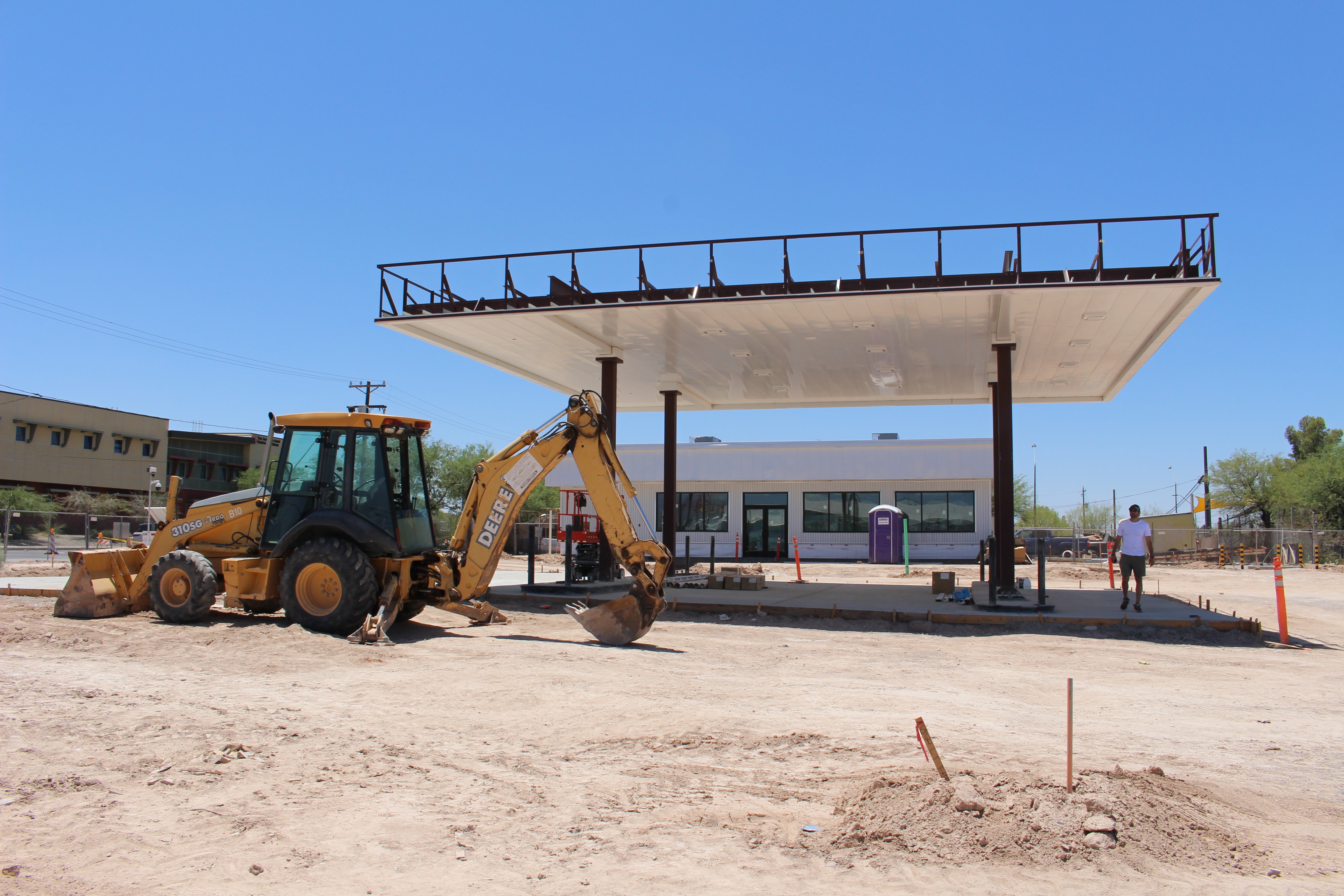 Construction site with a yellow backhoe loader in front of a building, under a large canopy structure, with a person nearby.
