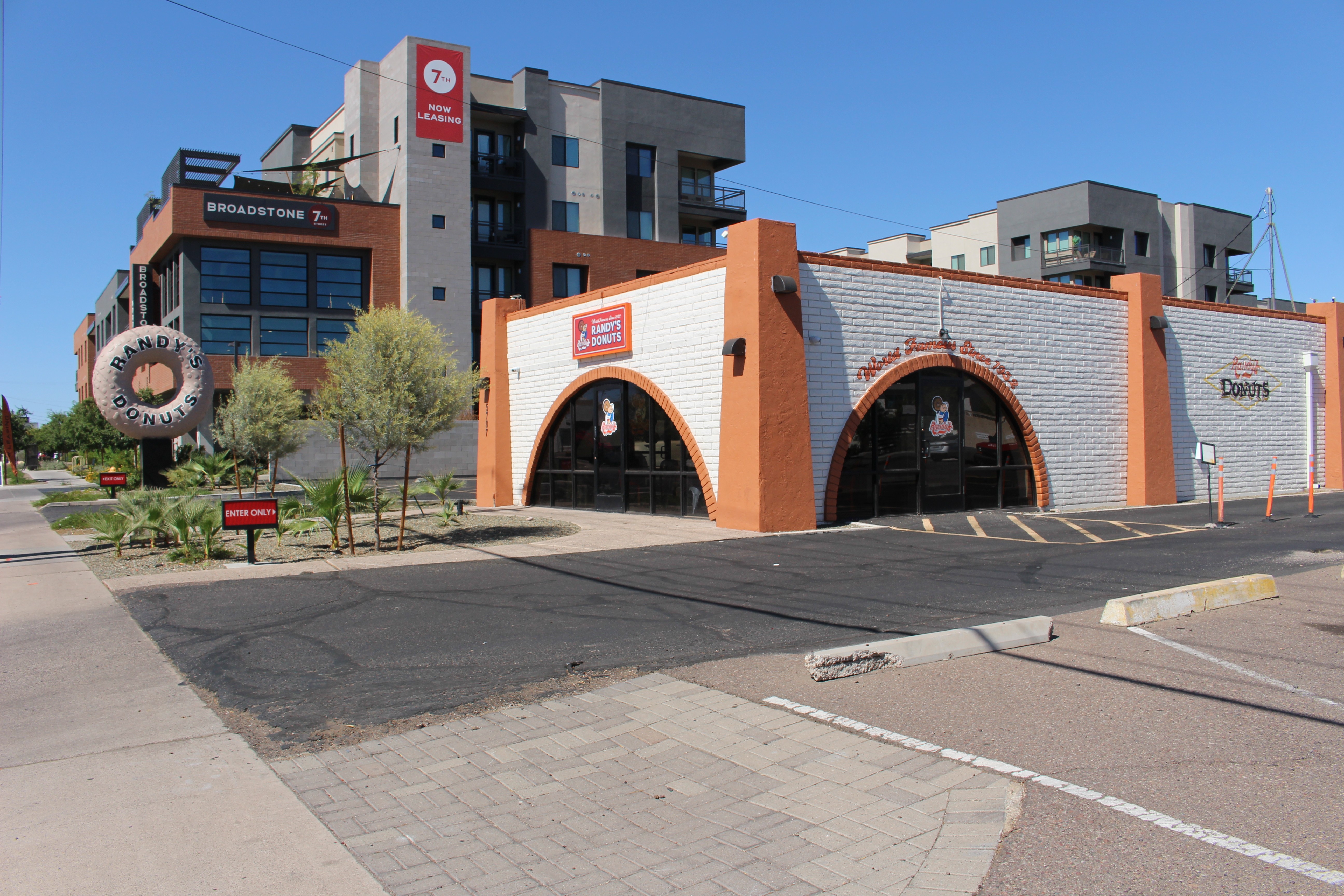 Commercial building with large windows and a parking lot in front, trees and a clear blue sky.