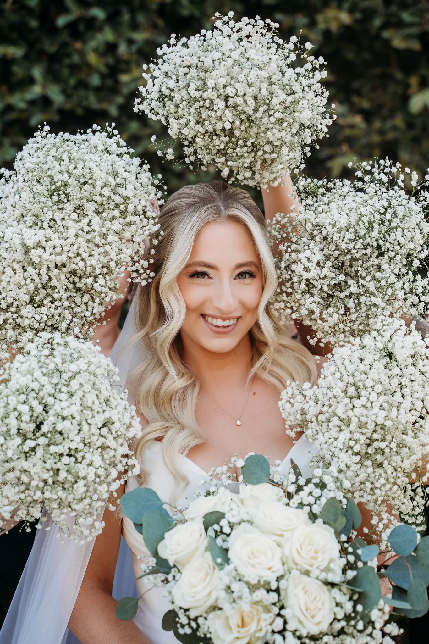 Smiling woman with long blonde hair surrounded by white flowers, holding a bouquet of white roses and greenery.