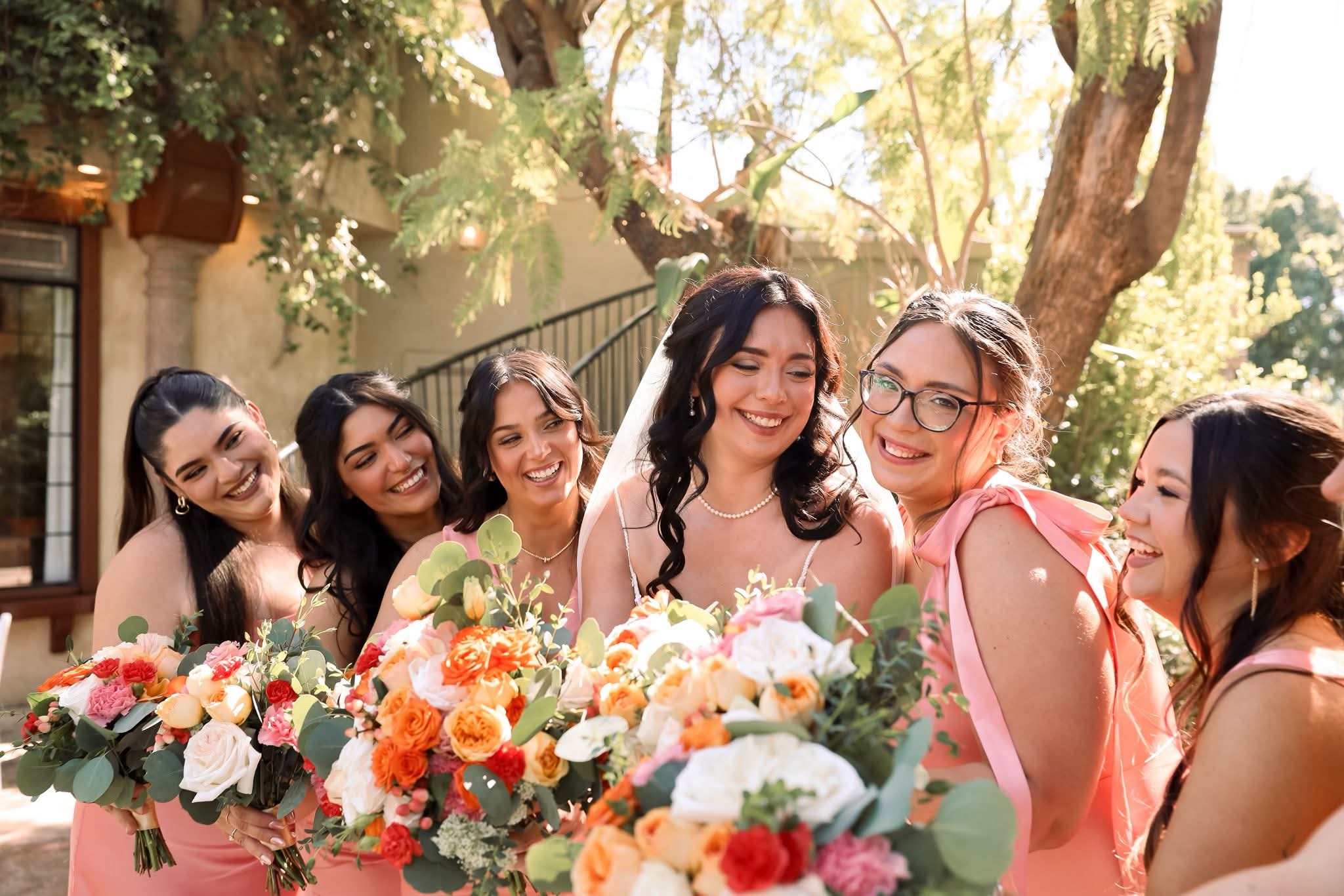 Group of six women smiling and holding bouquets outdoors with trees and sunlight in background.