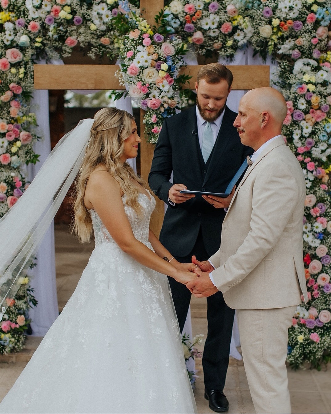 Bride and groom holding hands during wedding ceremony with officiant, surrounded by floral arch.