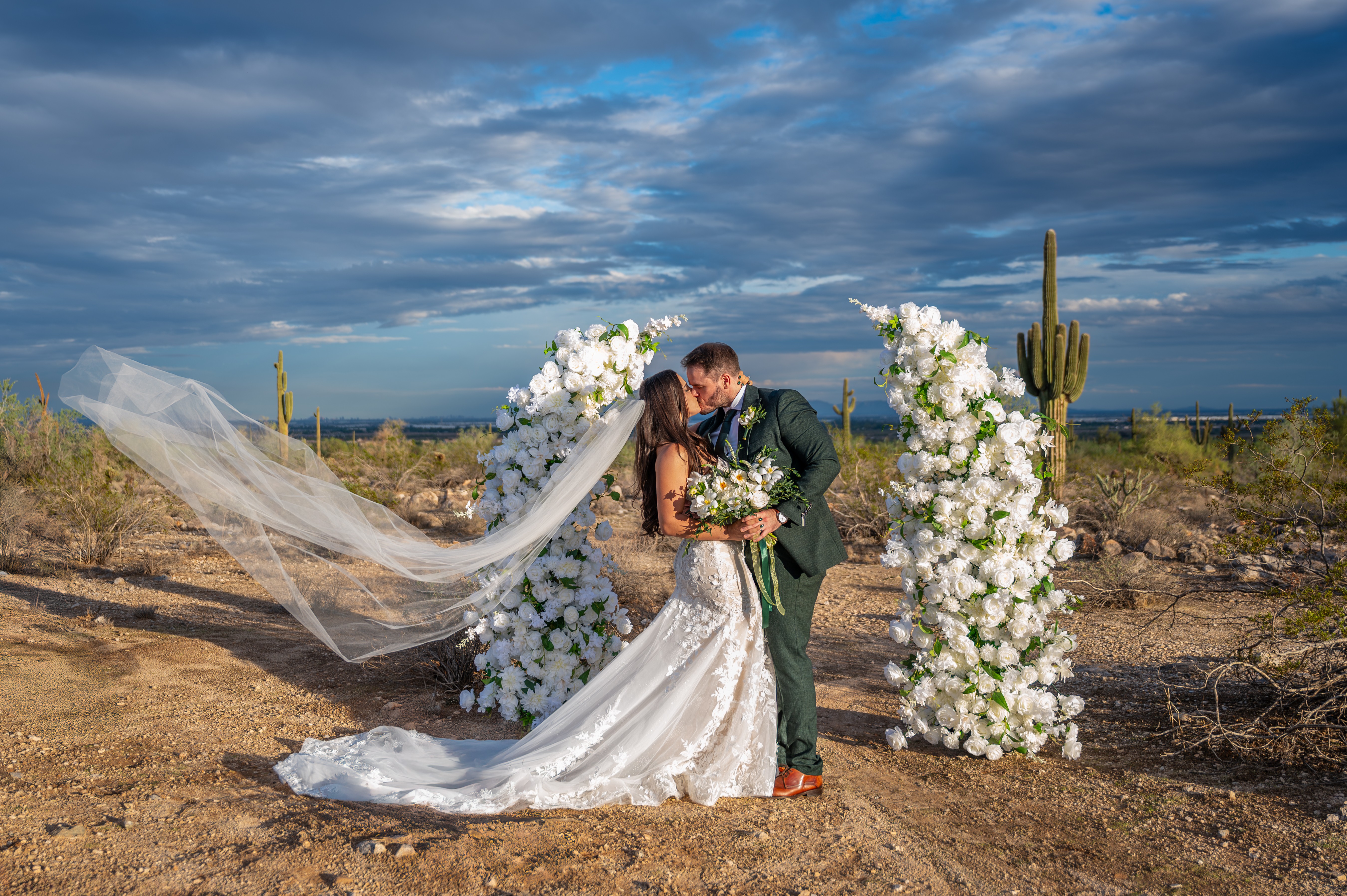 Bride and groom kissing outdoors with floral arch and desert landscape, cloudy sky in background.