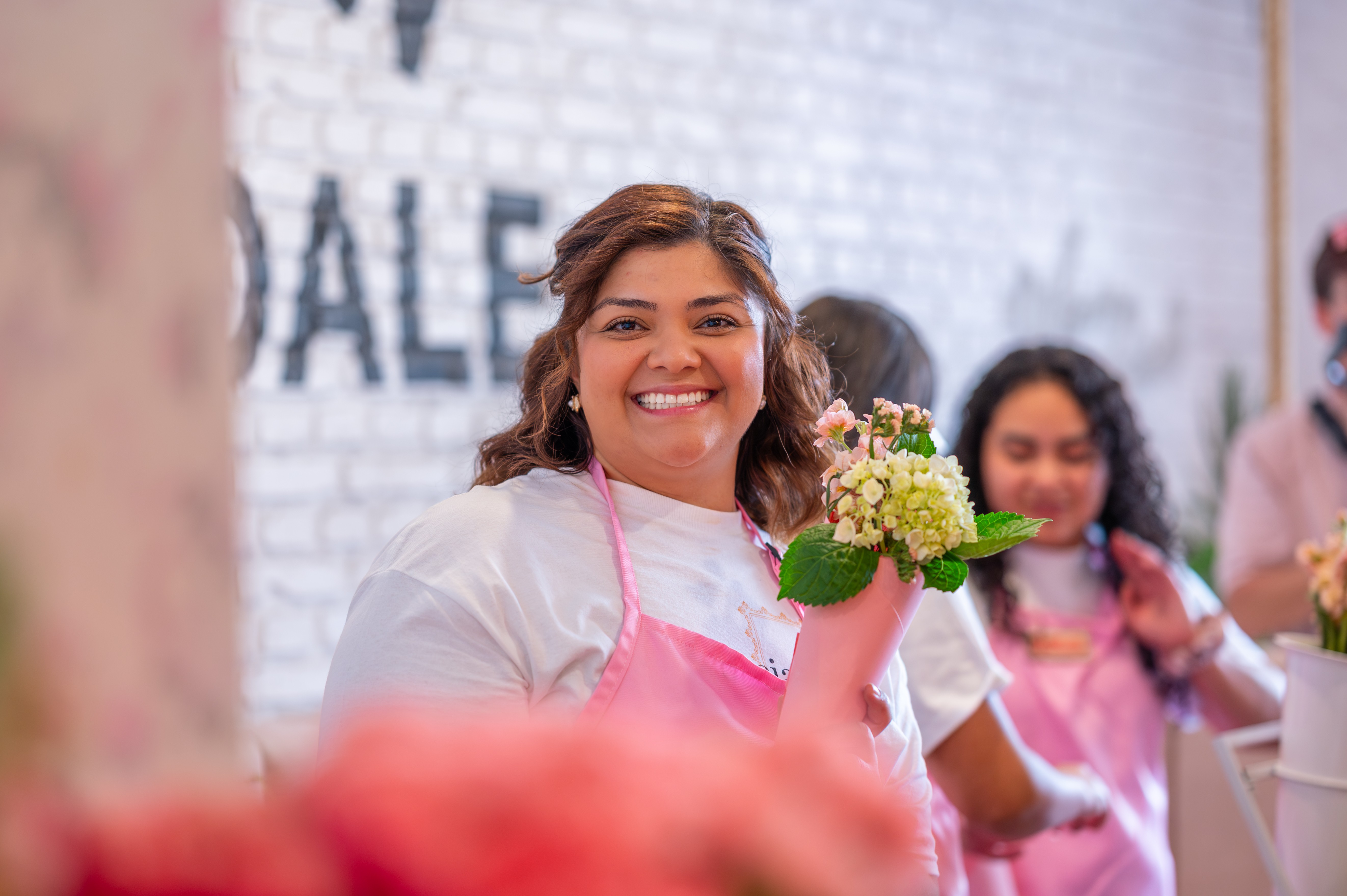 Smiling woman holding a small bouquet of flowers in a group setting, wearing a pink apron, with others in the background.
