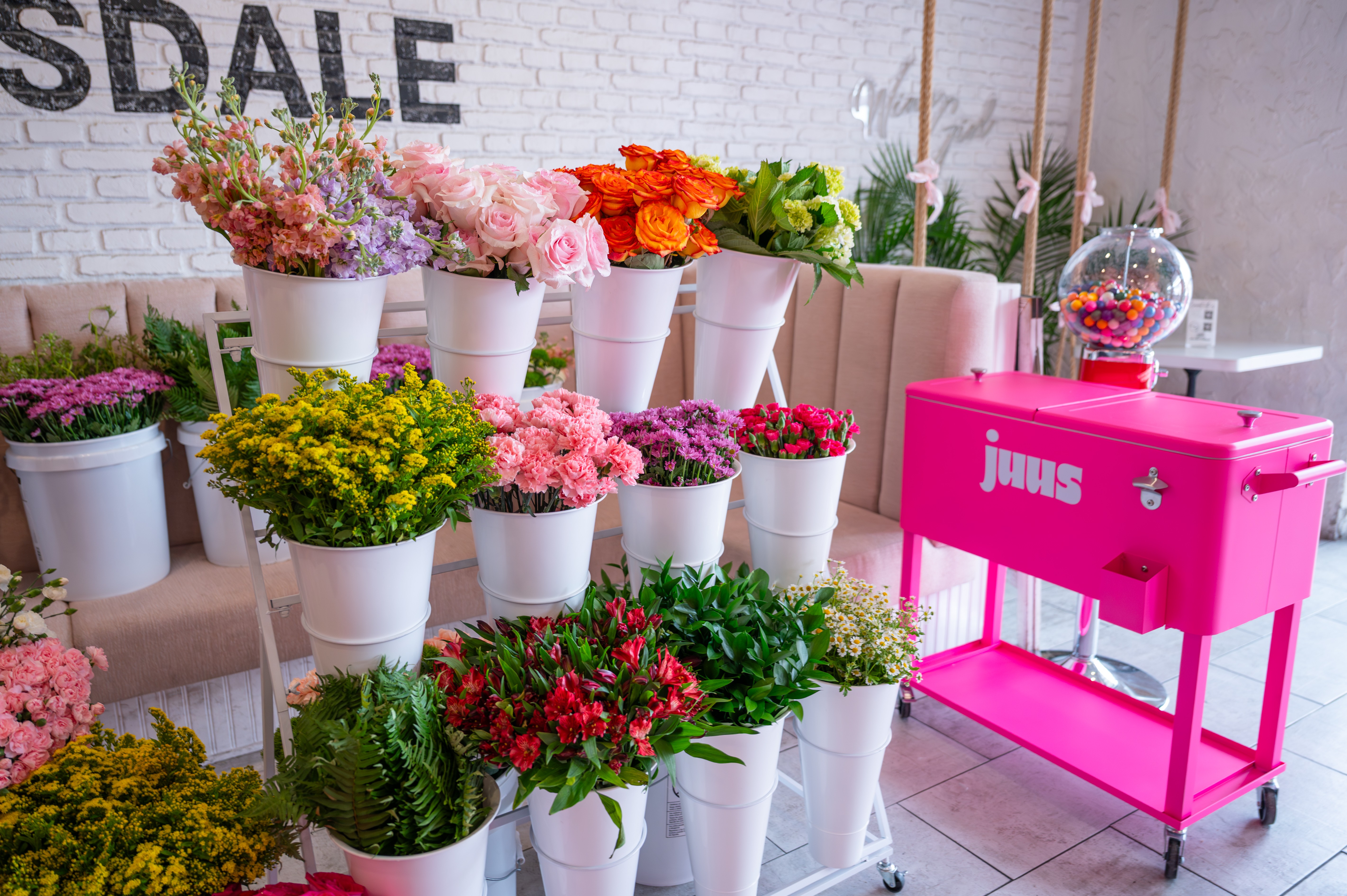 Display of colorful flowers in white pots and a bright pink Juras machine against a white brick wall, with a white sofa.