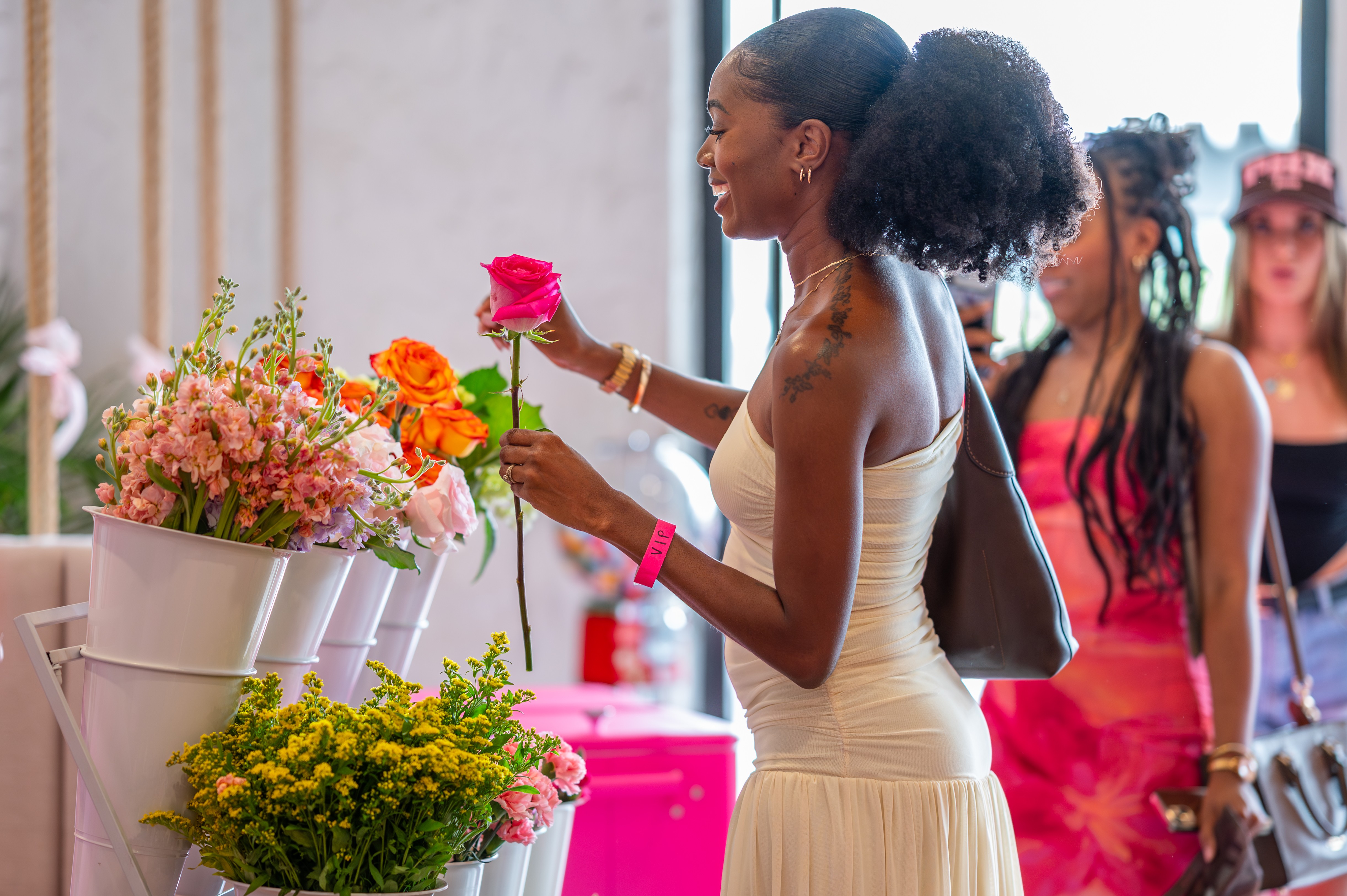 Woman in white dress holding pink and orange flowers, with other women in background, indoors with large windows