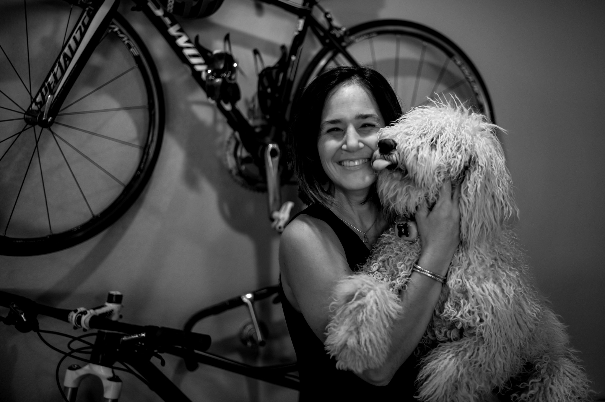Woman smiling while holding a fluffy dog indoors, with bicycles mounted on the wall behind her.