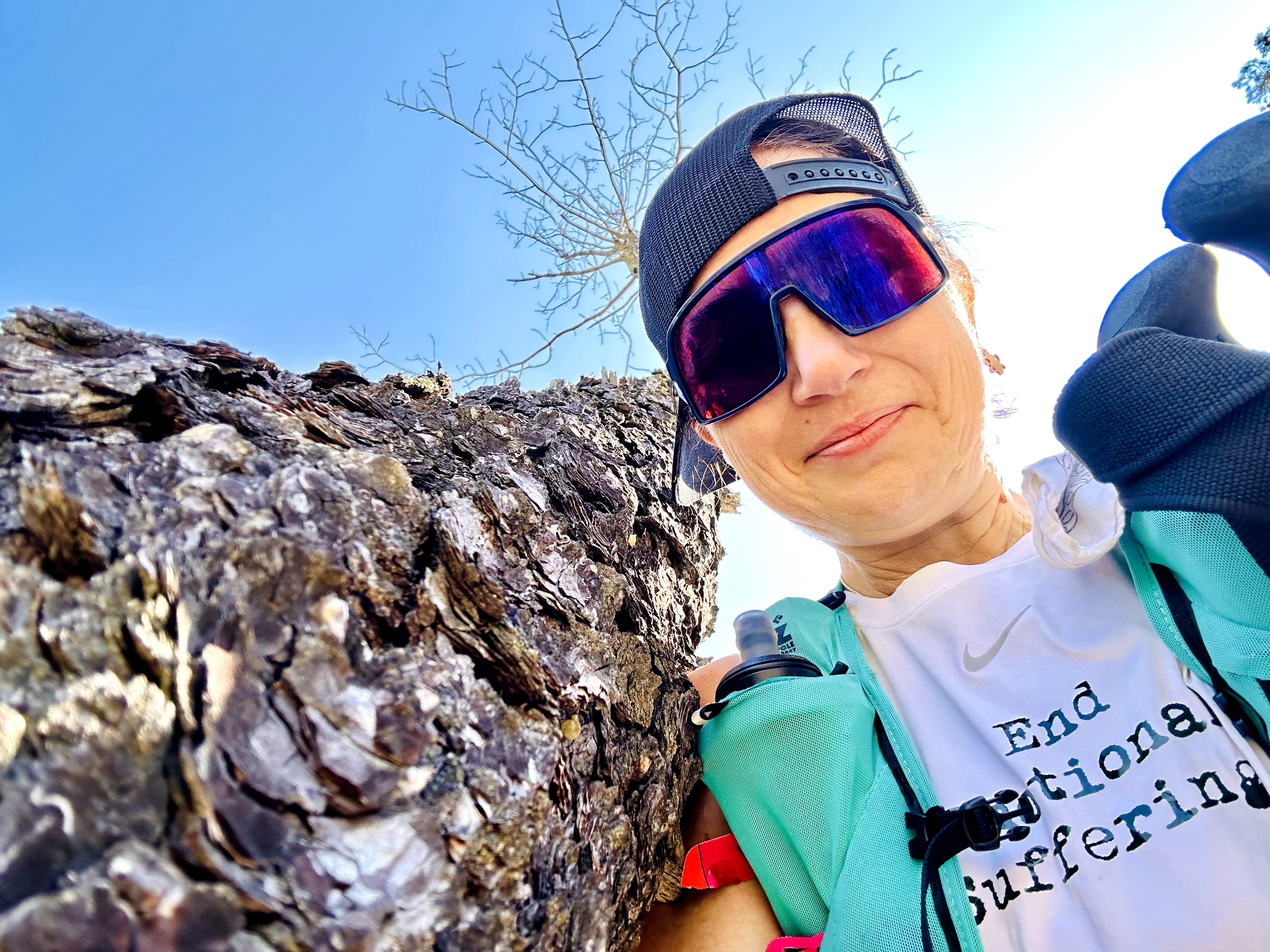 Person outdoors wearing sunglasses, cap, and sports gear, smiling near a tree and rocky surface.