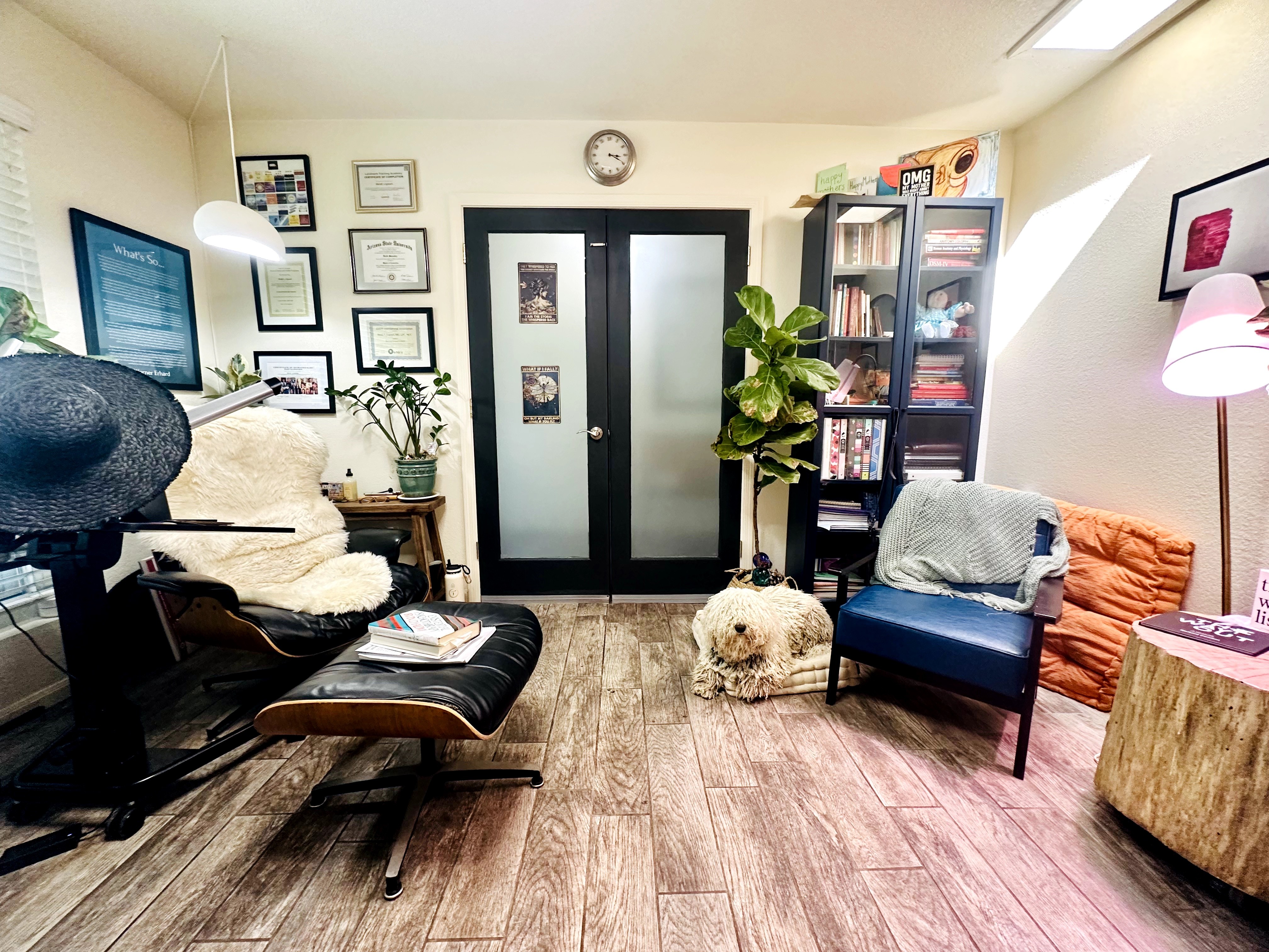 Living room with chairs, bookshelf, plants, and wall decorations, illuminated by natural and artificial light.