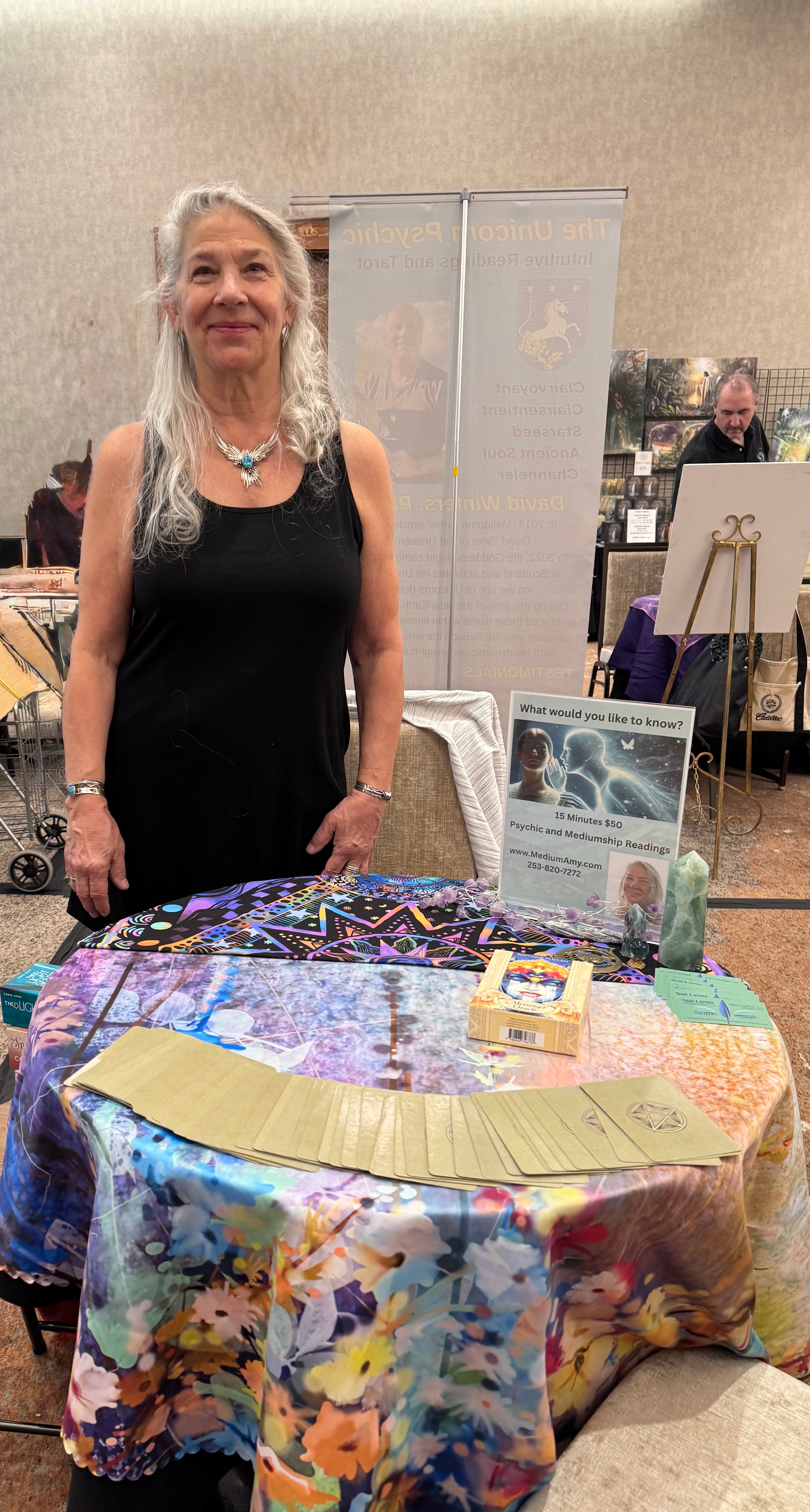 Woman with long gray hair standing behind a table with colorful cloth, in an indoor event setting.