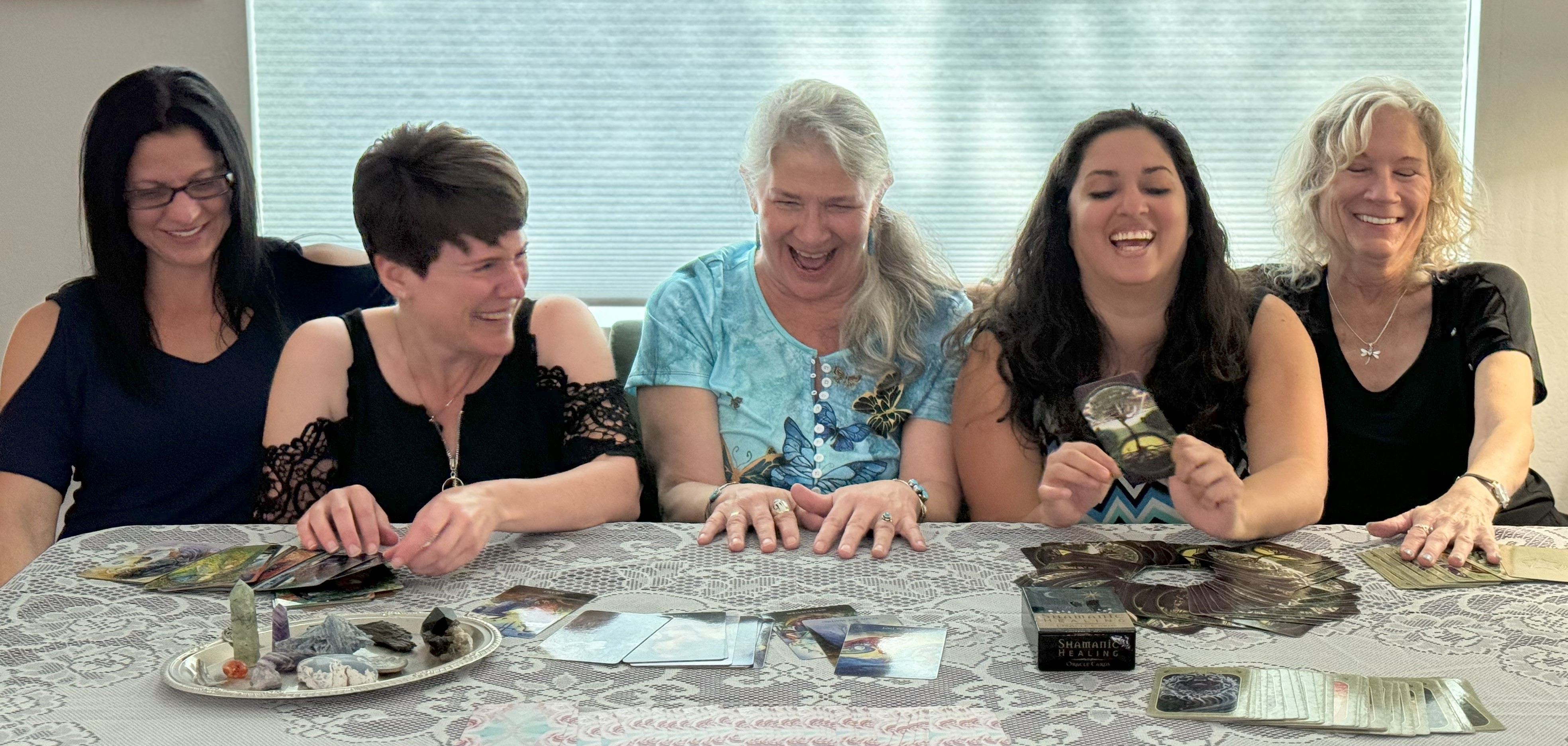 Five women sitting at a table, smiling and laughing, with cards and snacks on the table.