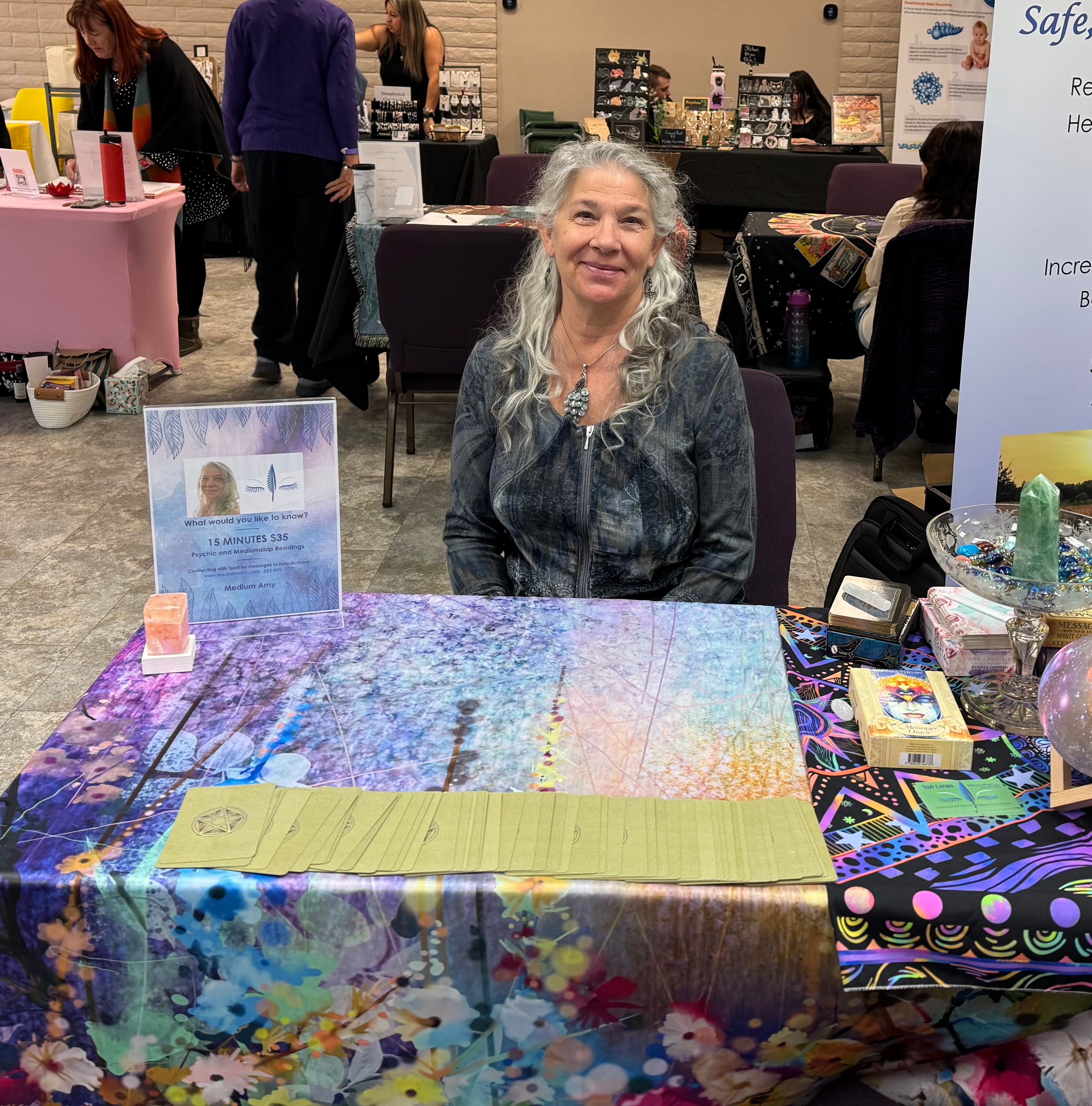 Smiling woman with long gray hair sitting at a colorful table in an indoor event space.