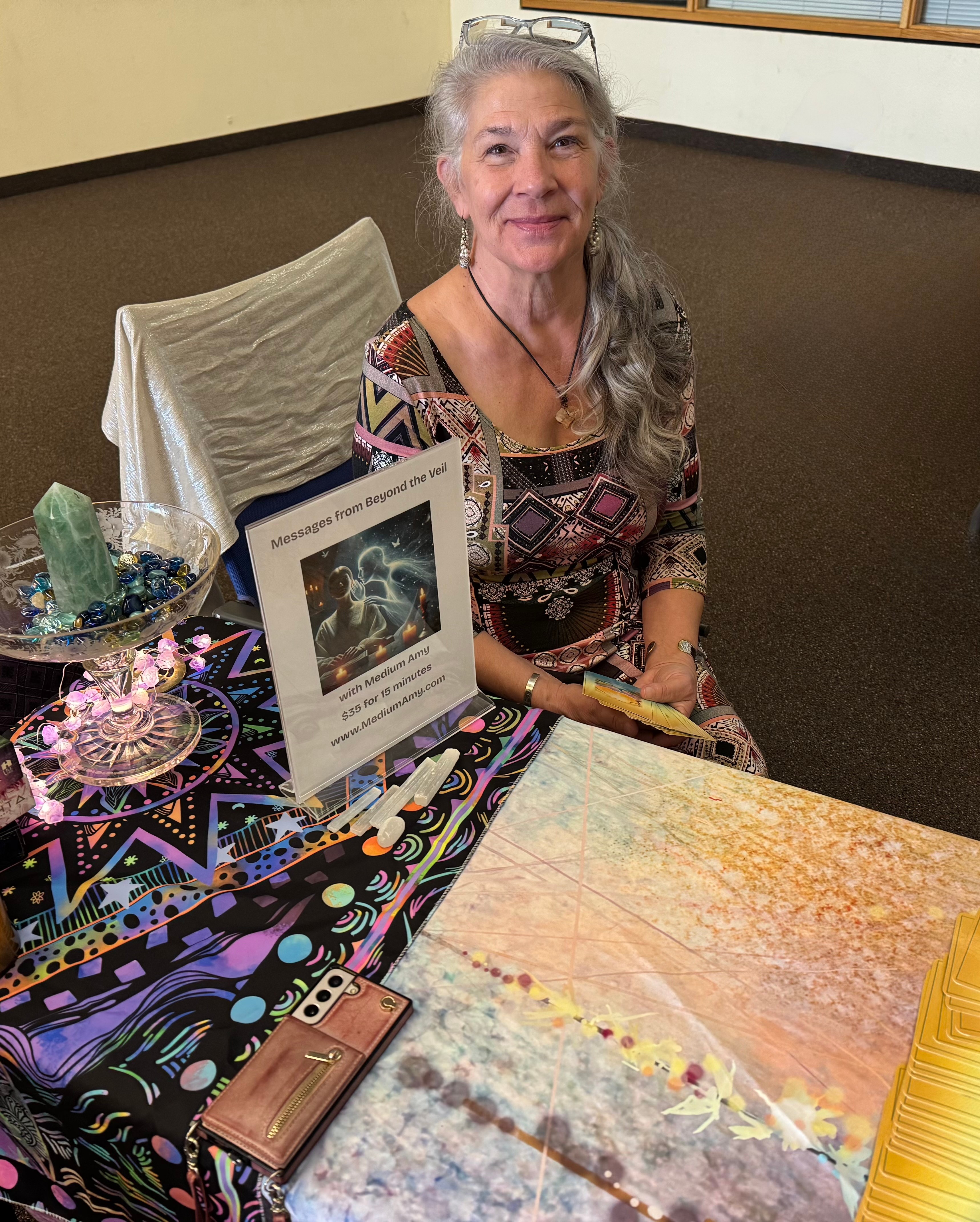 Older woman with long gray hair sitting at a table with colorful decorations and a sign, smiling at the camera.