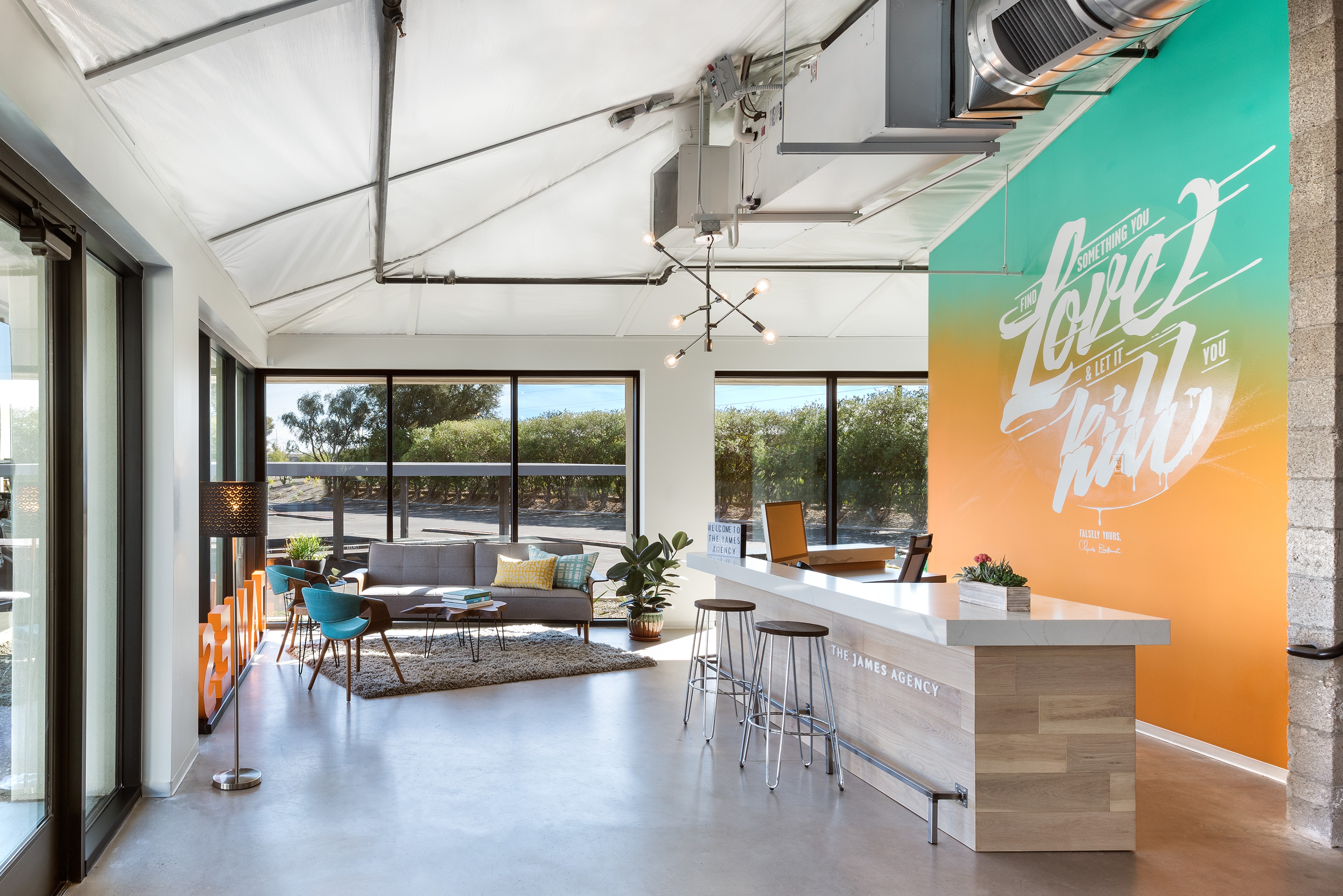 Modern interior with large windows, a seating area, a bar counter with stools, and a colorful wall with text.