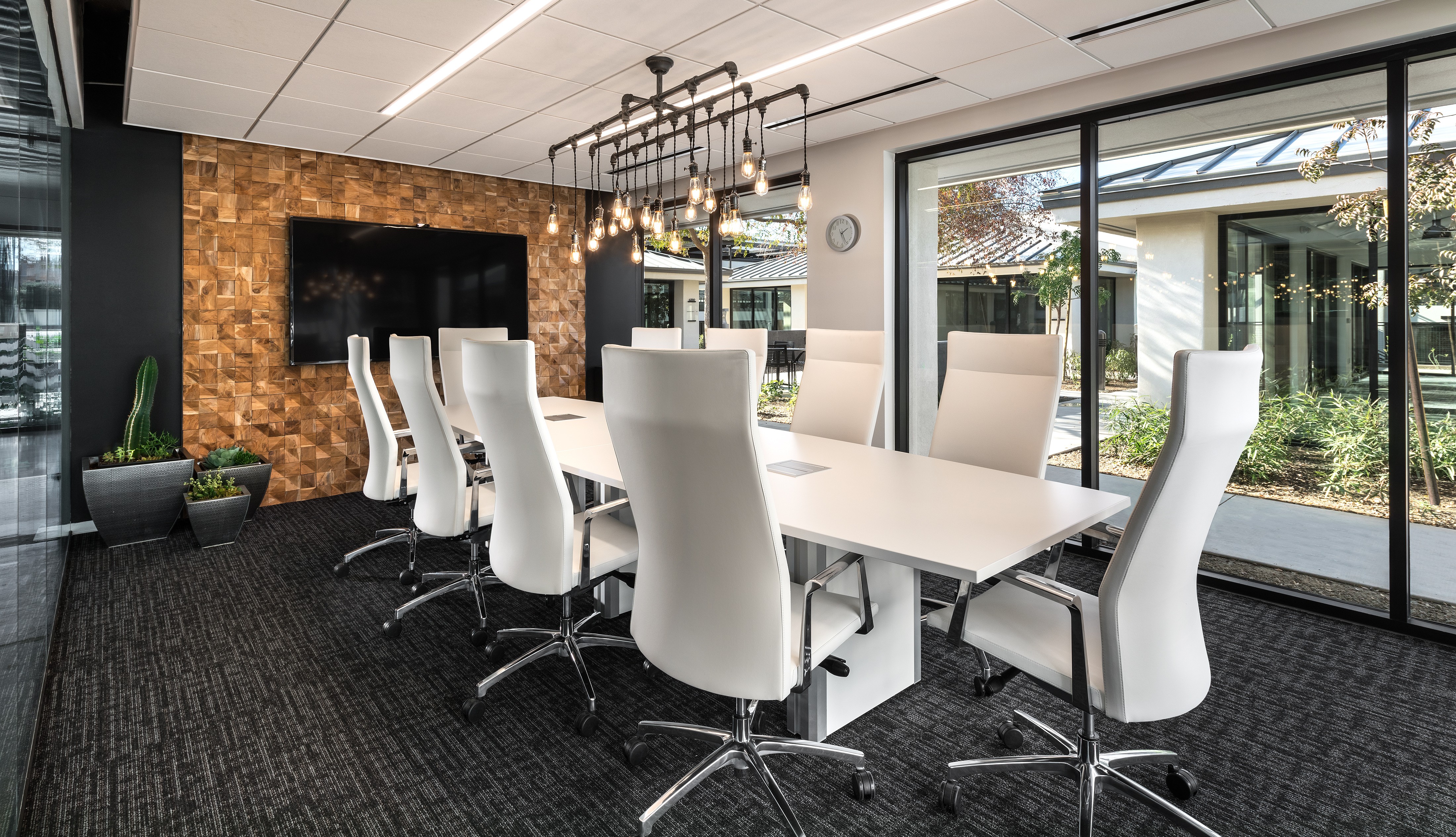 Modern conference room with white chairs around a long table, large windows, and a brick accent wall with a TV.