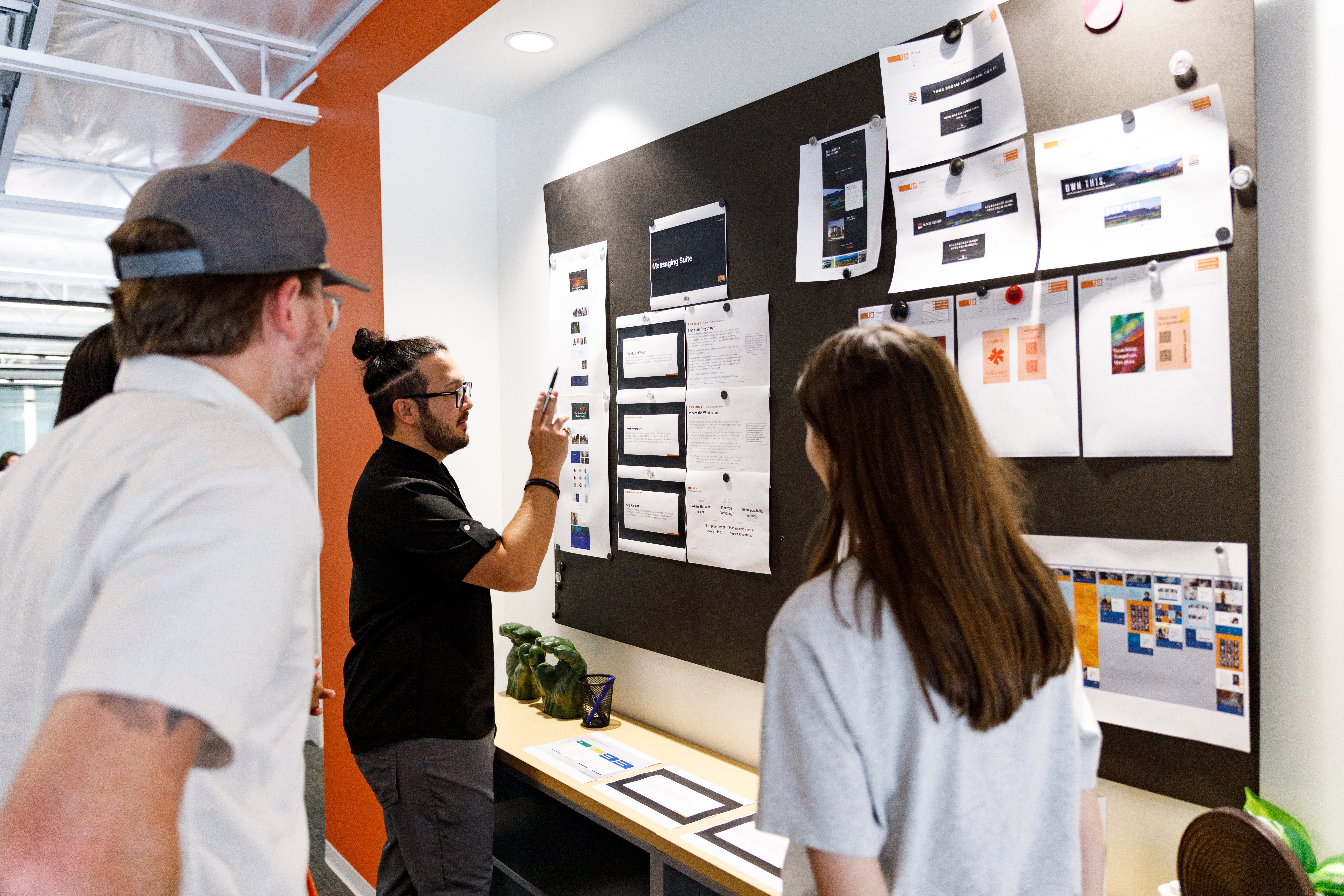 Three people in a meeting room looking at a blackboard with papers and charts attached, discussing ideas.