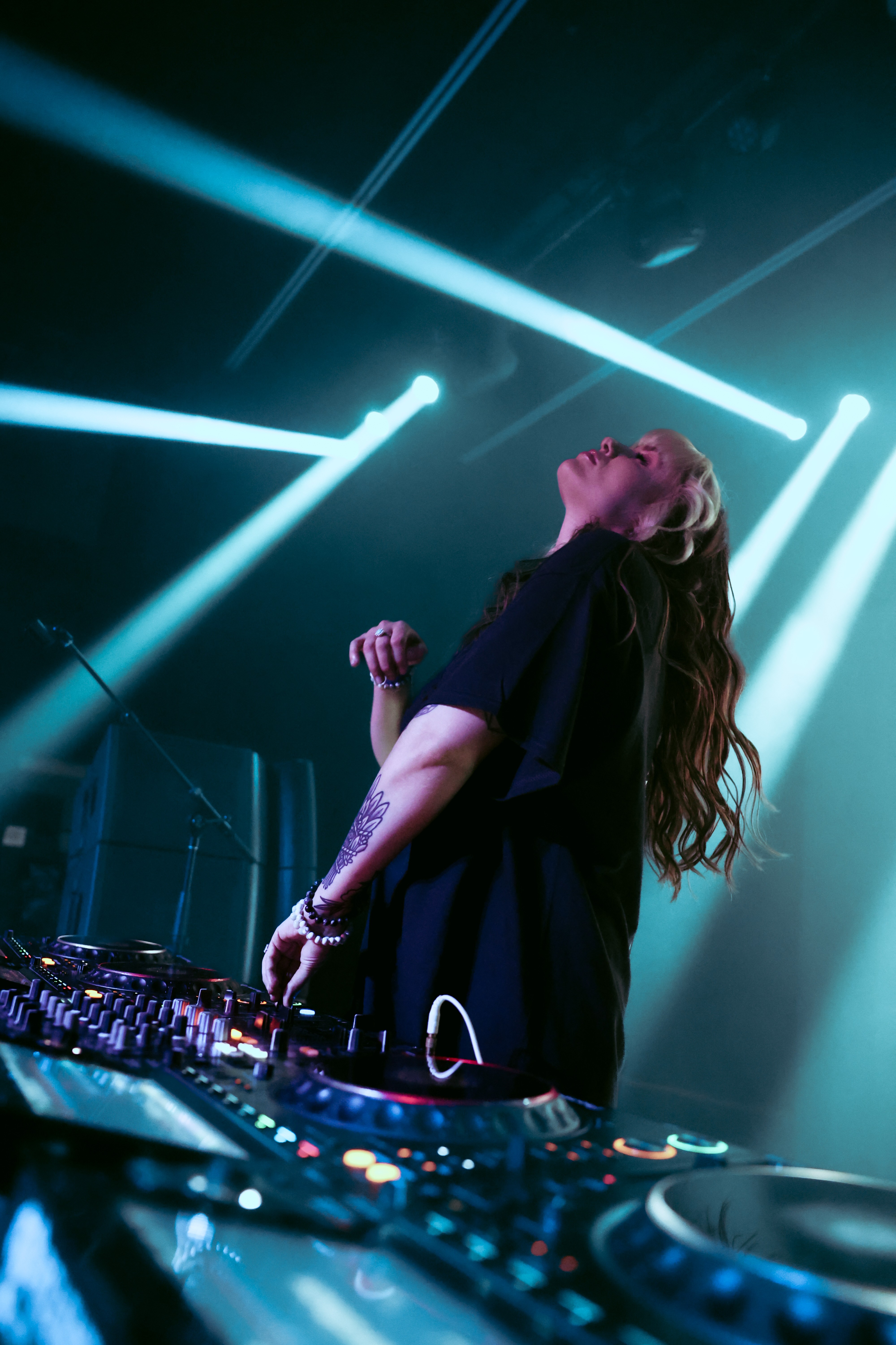 DJ with long hair performing on stage with bright blue and white lights shining behind her.
