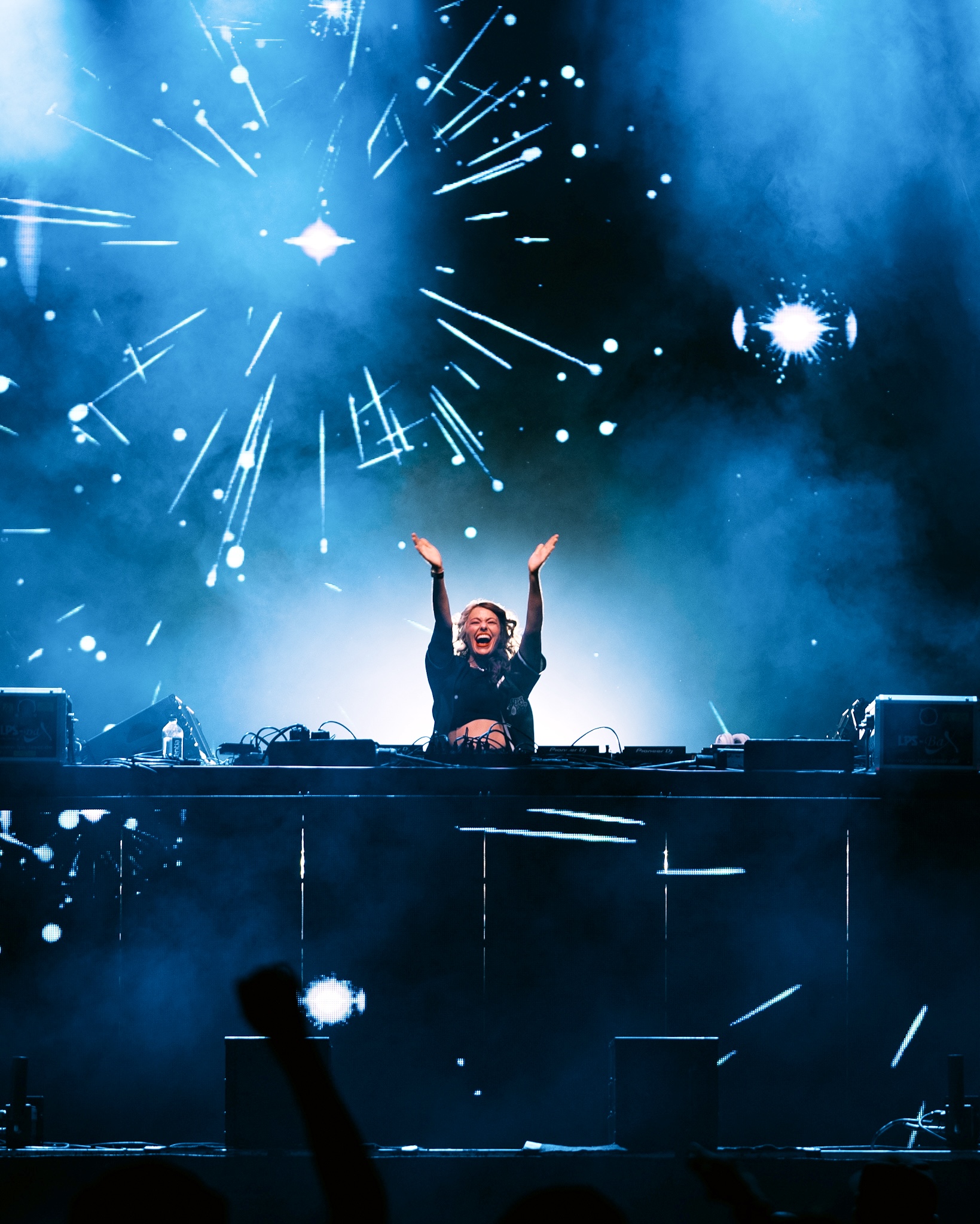 DJ with arms raised performing on stage with fireworks and blue lighting, crowd visible in foreground.