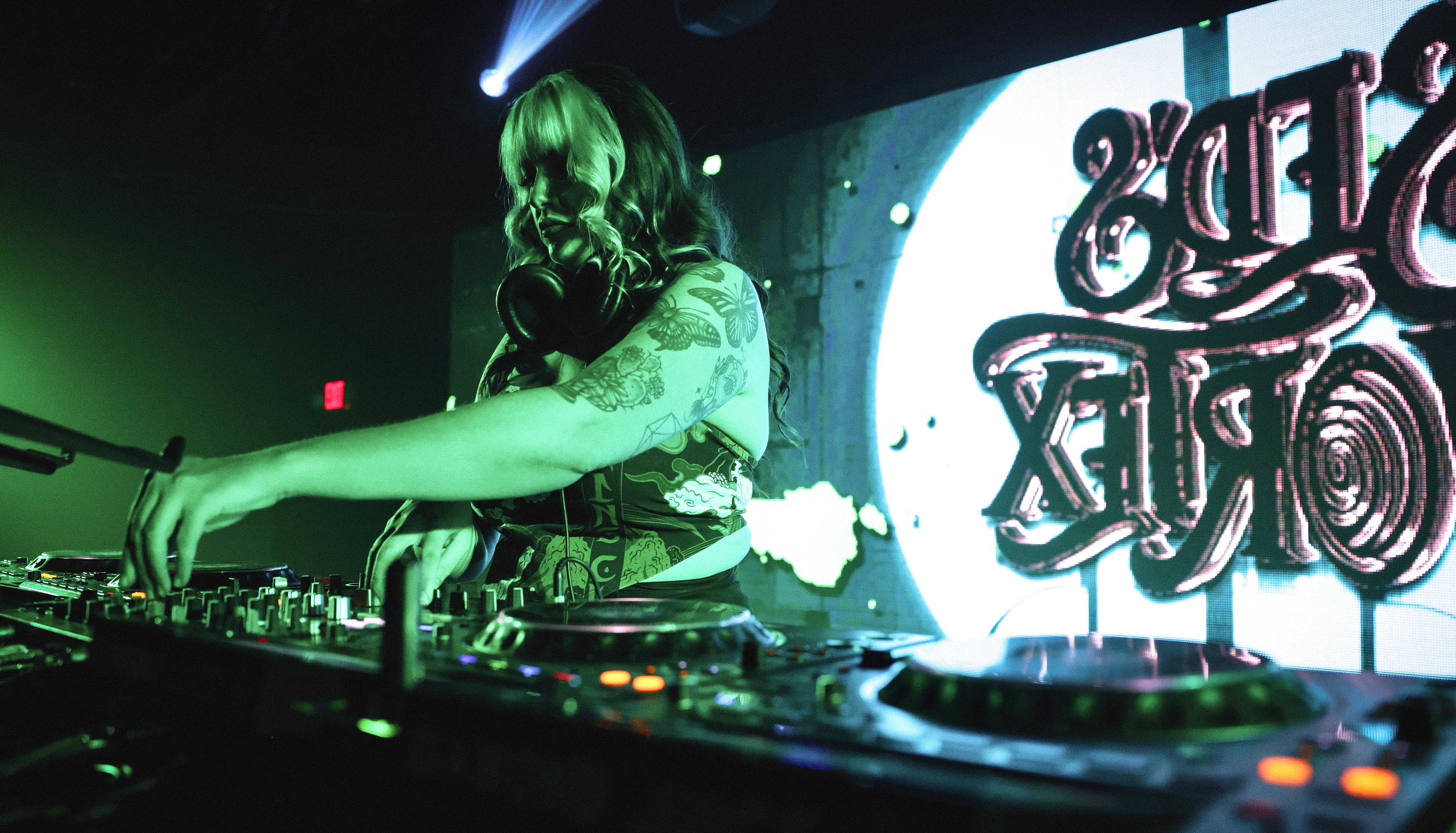 DJ with long hair operating turntables on stage with a large screen displaying text behind her.