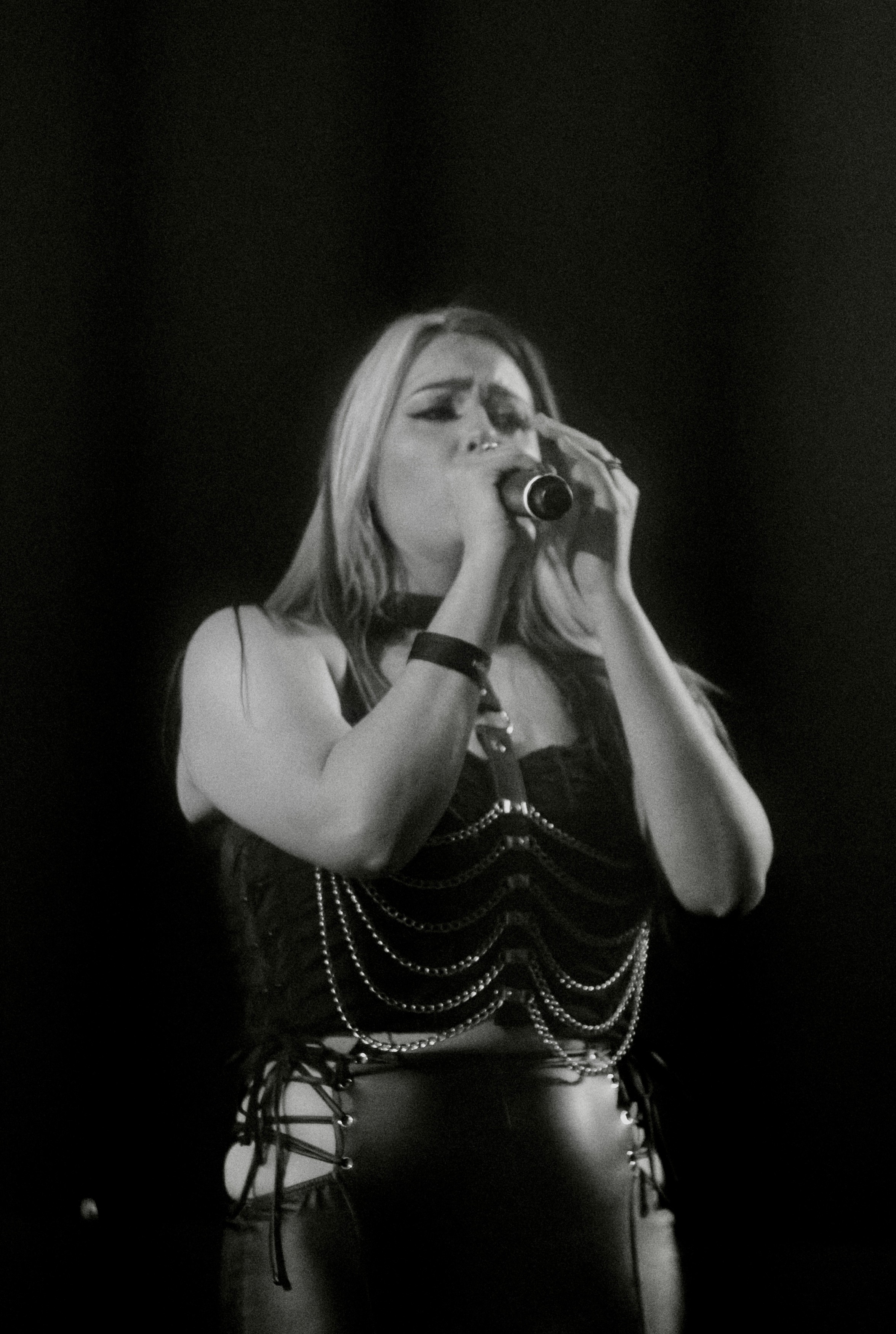 A woman singing into a microphone on stage, wearing a black outfit with chains, with dark background.