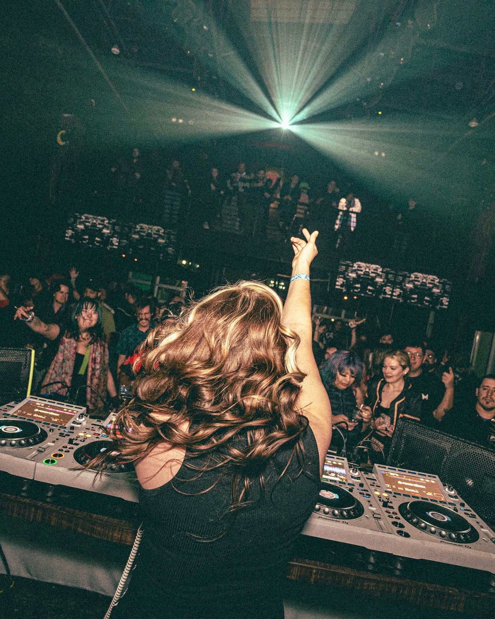 DJ with long wavy hair raising hand in a nightclub with a crowd and laser lights.