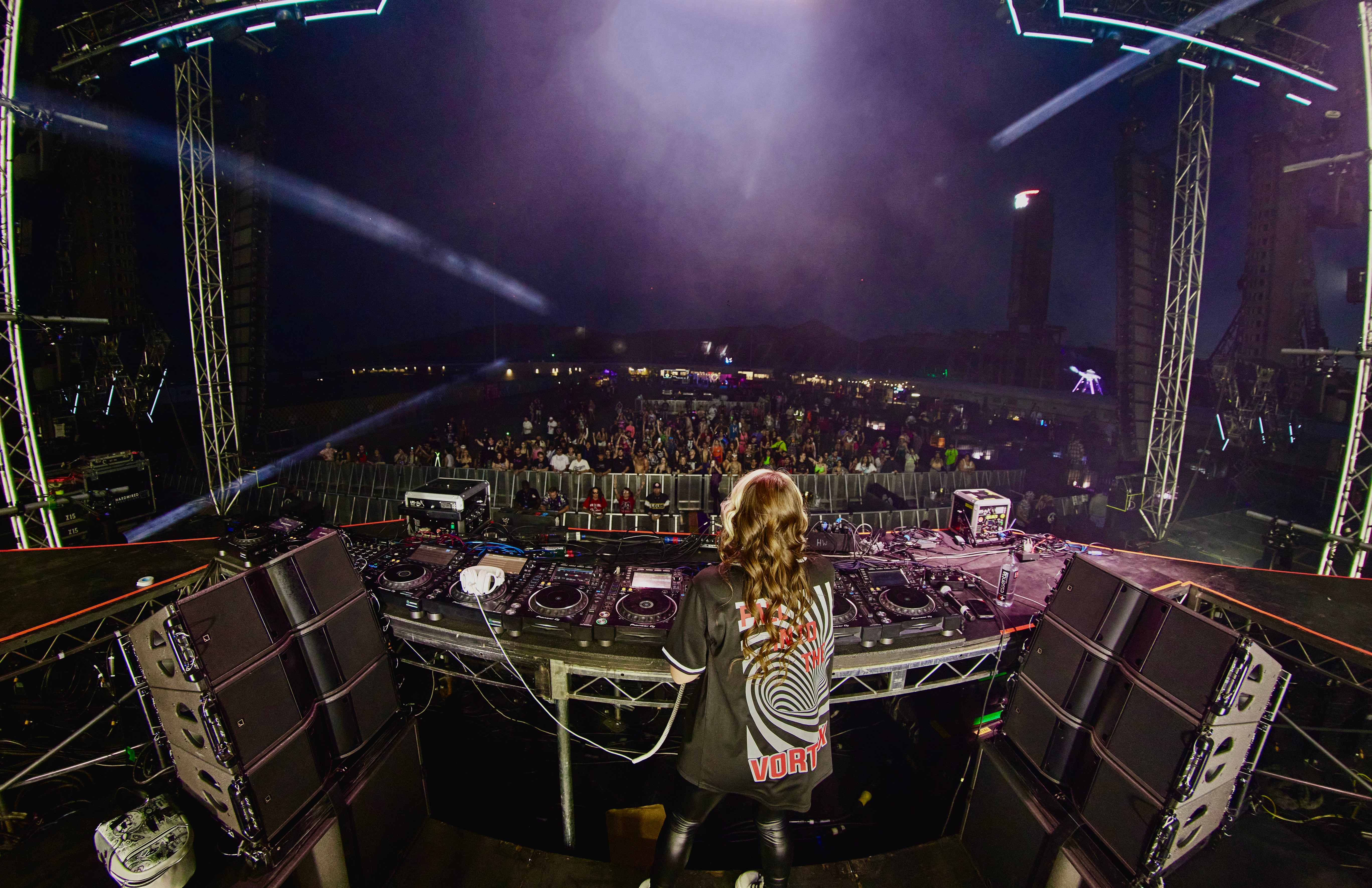 DJ performing at a nightclub with a large crowd, DJ equipment, and stage lighting, viewed from behind the DJ booth.