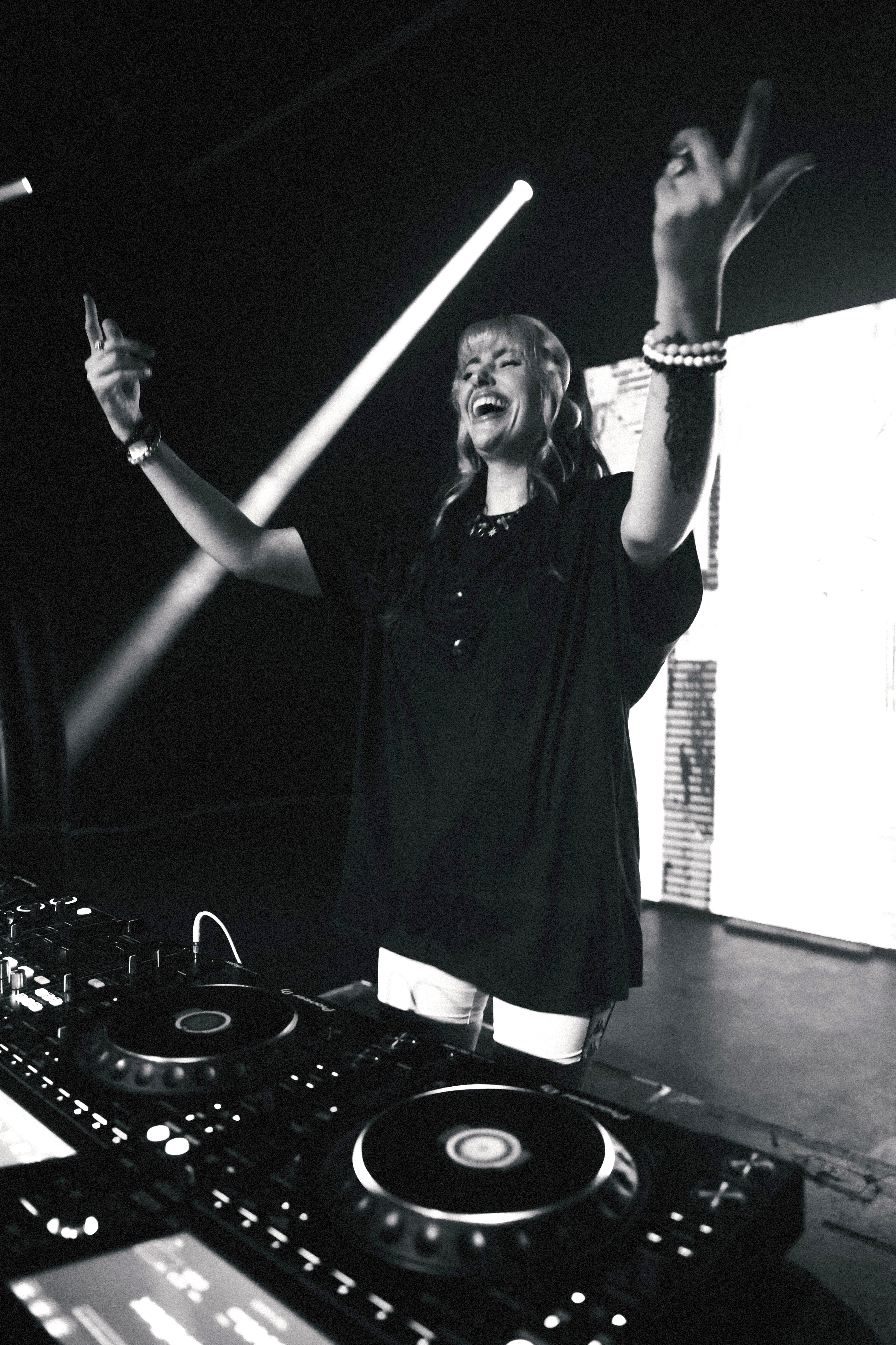 Woman DJ with arms raised, smiling, standing behind DJ equipment on stage, with a bright light and screen in background.