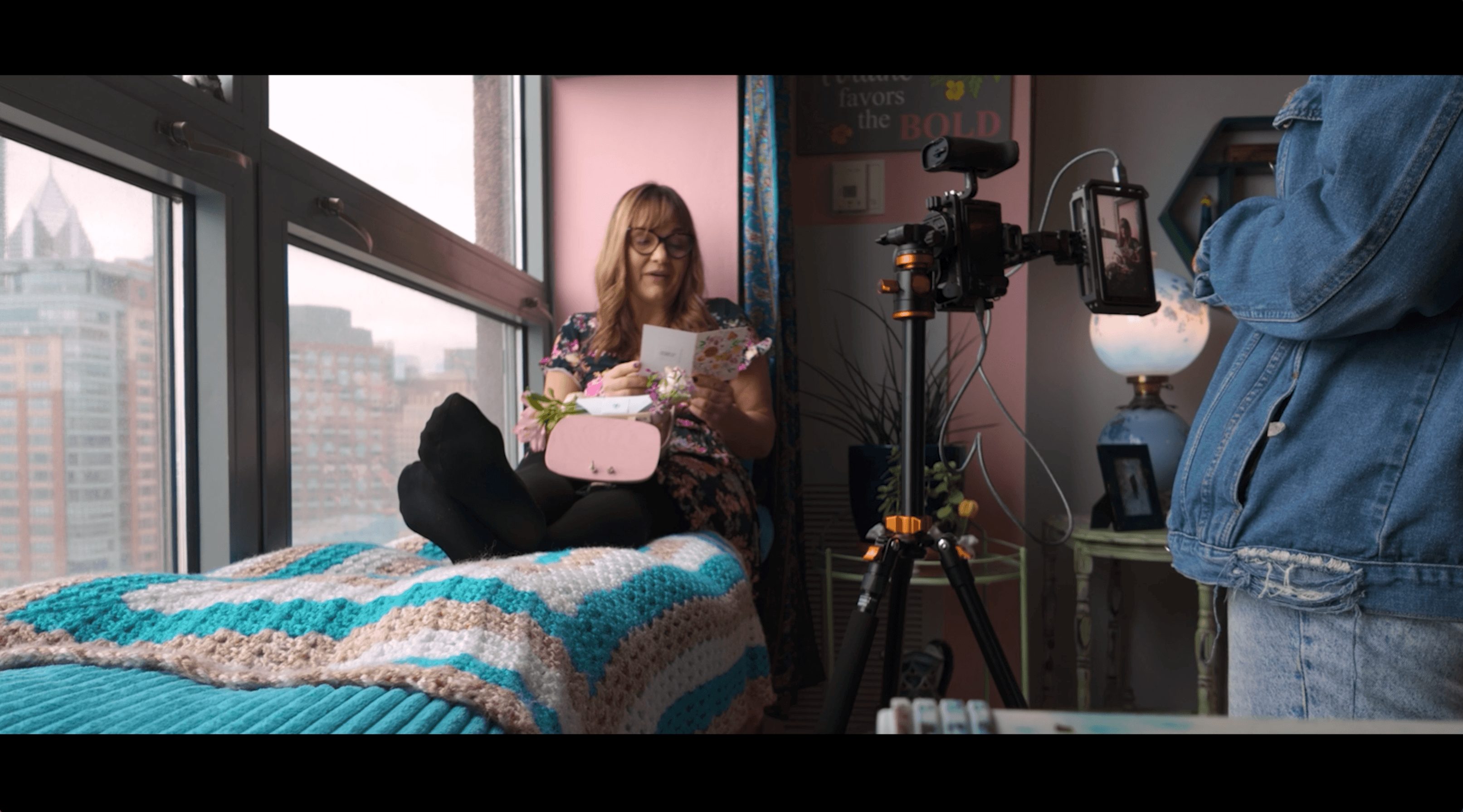 Person sitting on bed reading a book, camera operator filming in a room with large windows and city view
