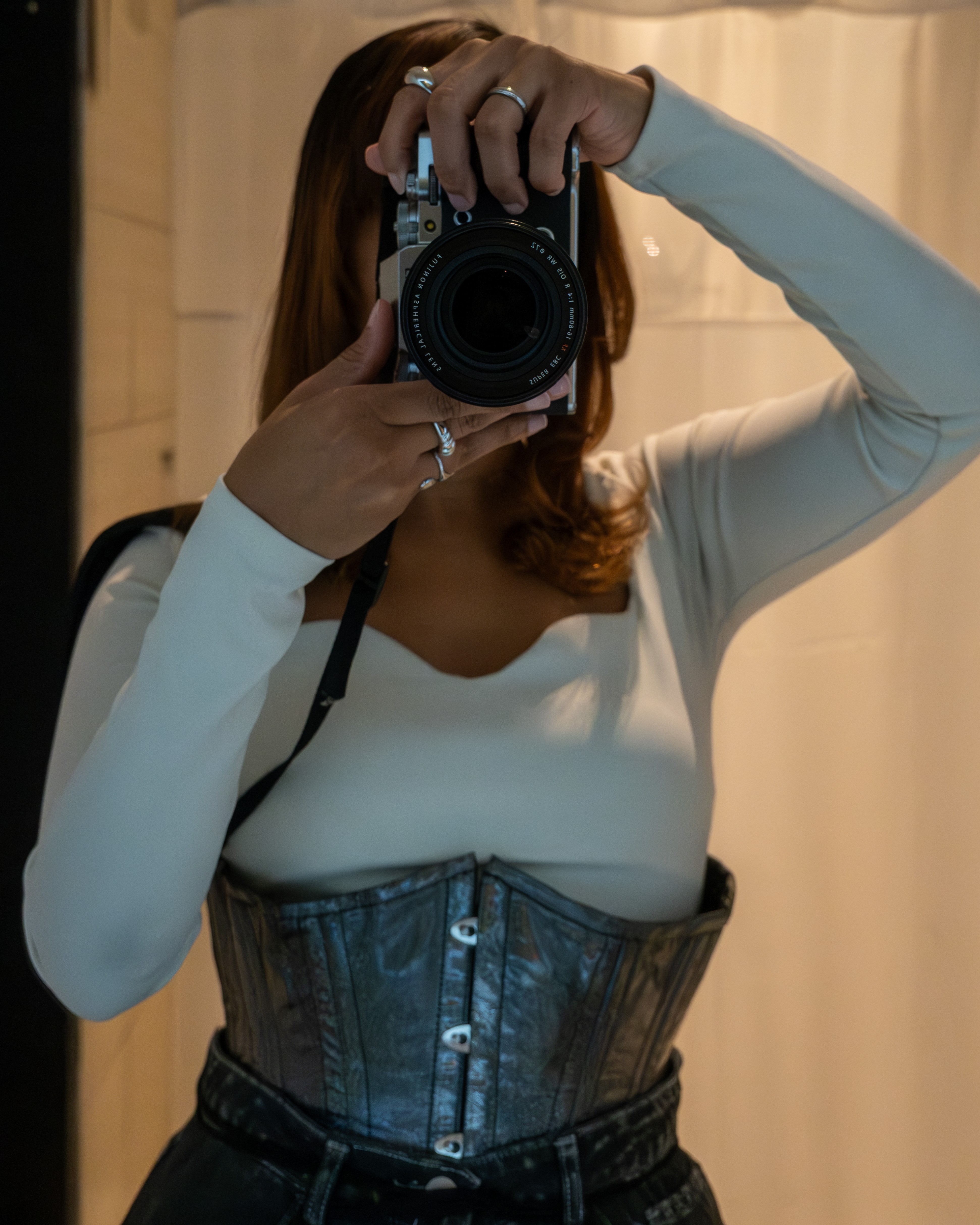 Person taking a mirror selfie with a camera, wearing a white top and a corset, with a curtain background.