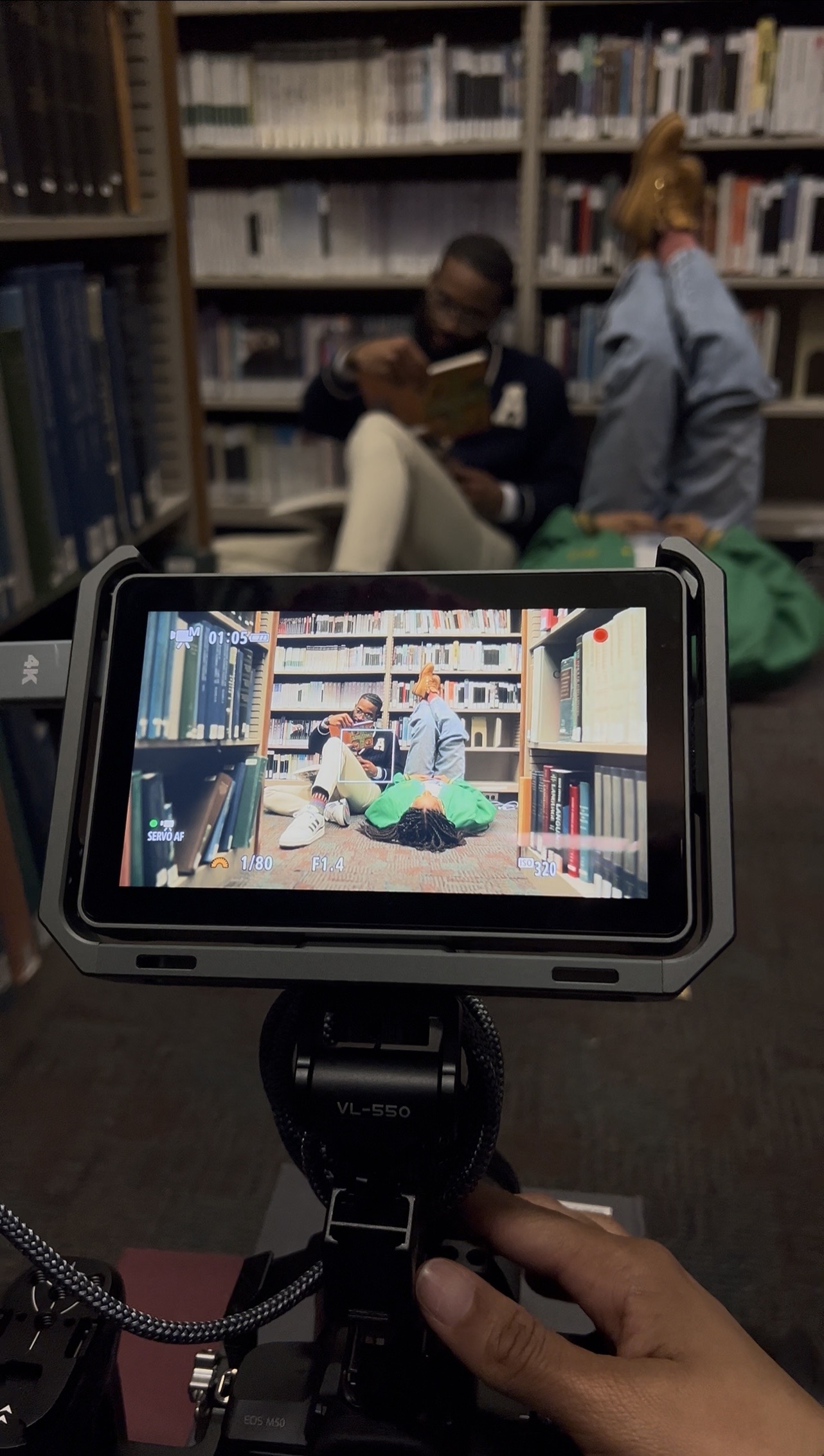 Person filming a scene with two people in a library, one reading a book, shelves in background.