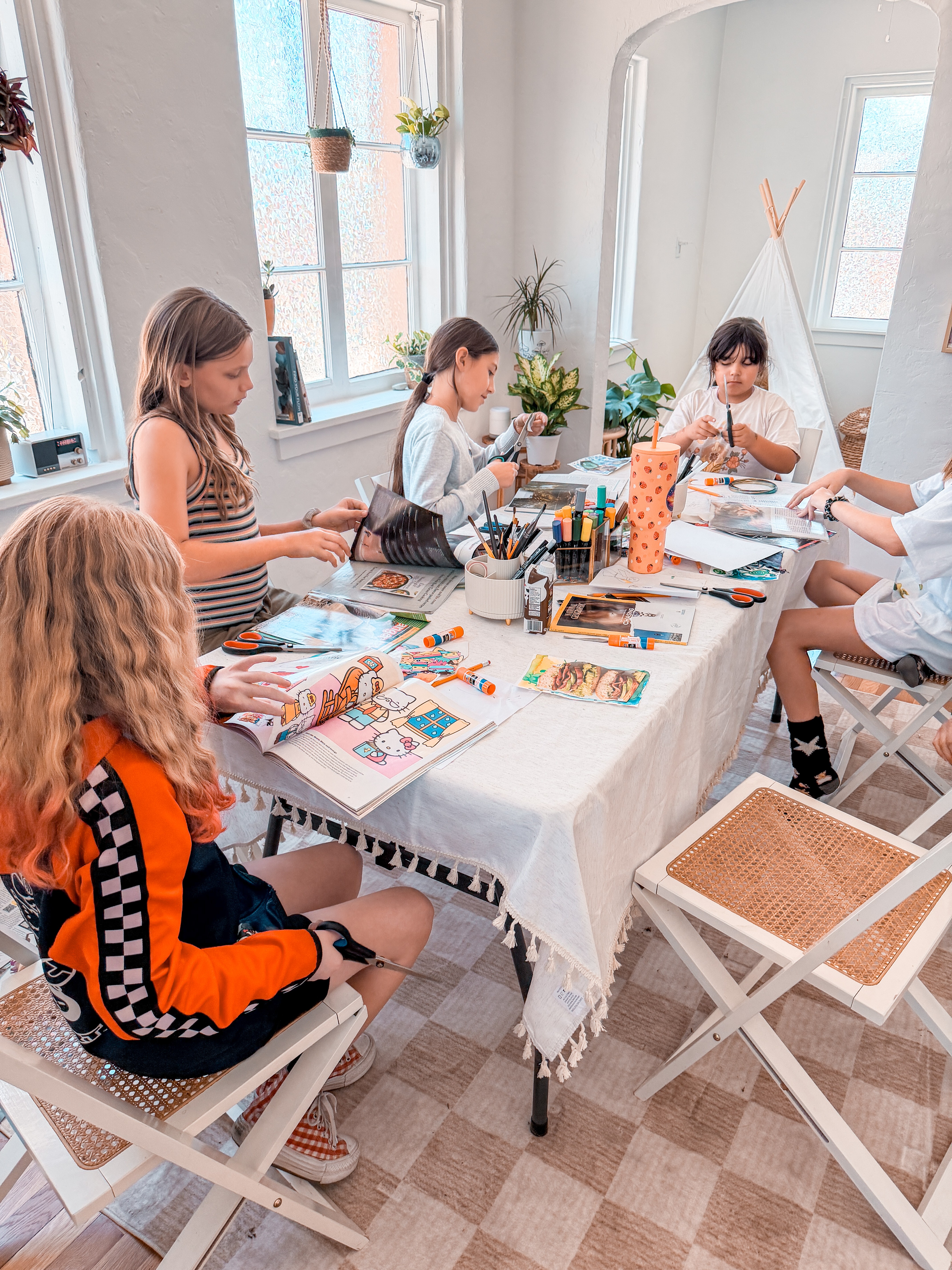 Children sit around a table engaged in arts and crafts, with windows and plants in the background.