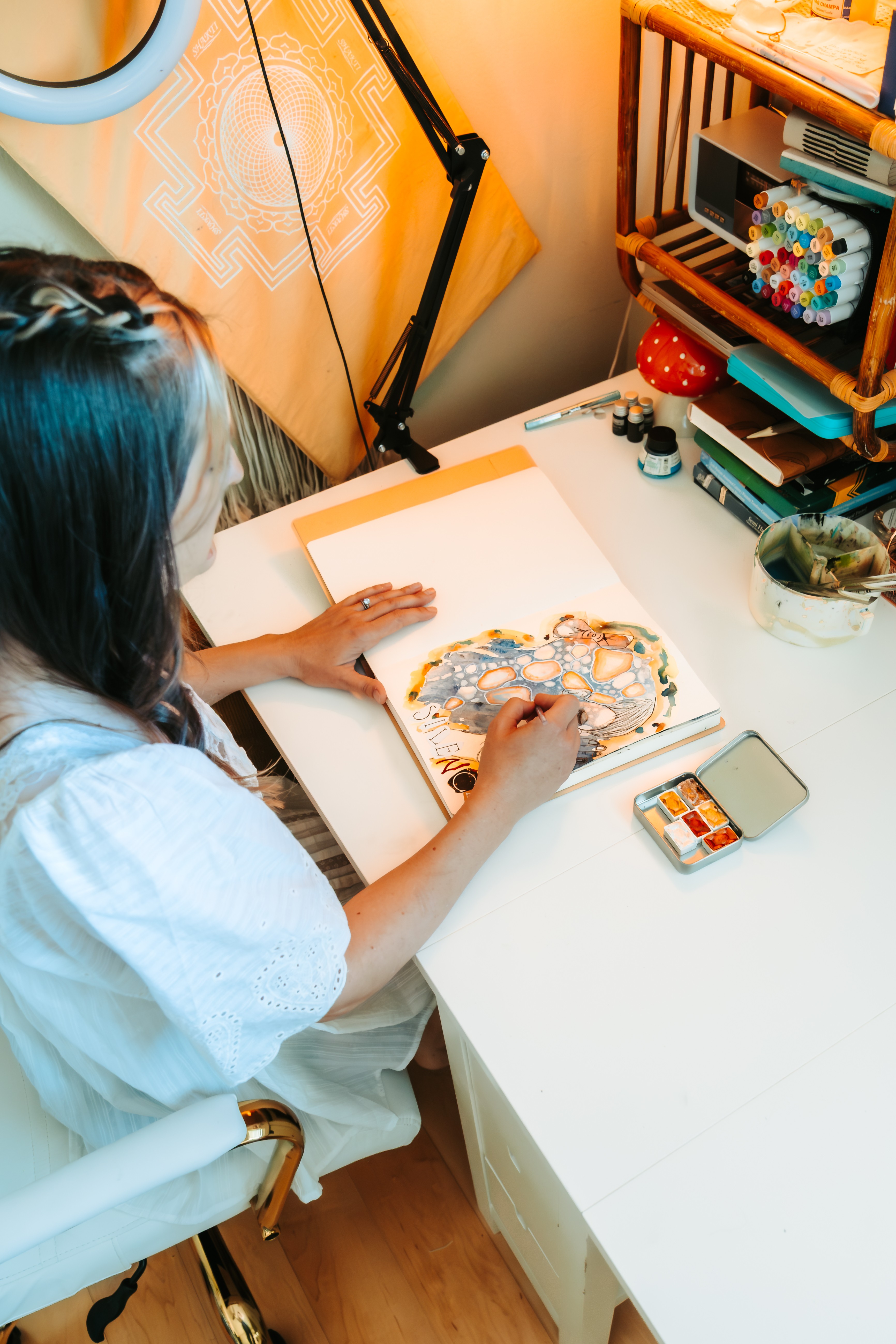 Person sitting at a desk drawing with colorful markers, with art supplies and a smartphone nearby, in a cozy room.