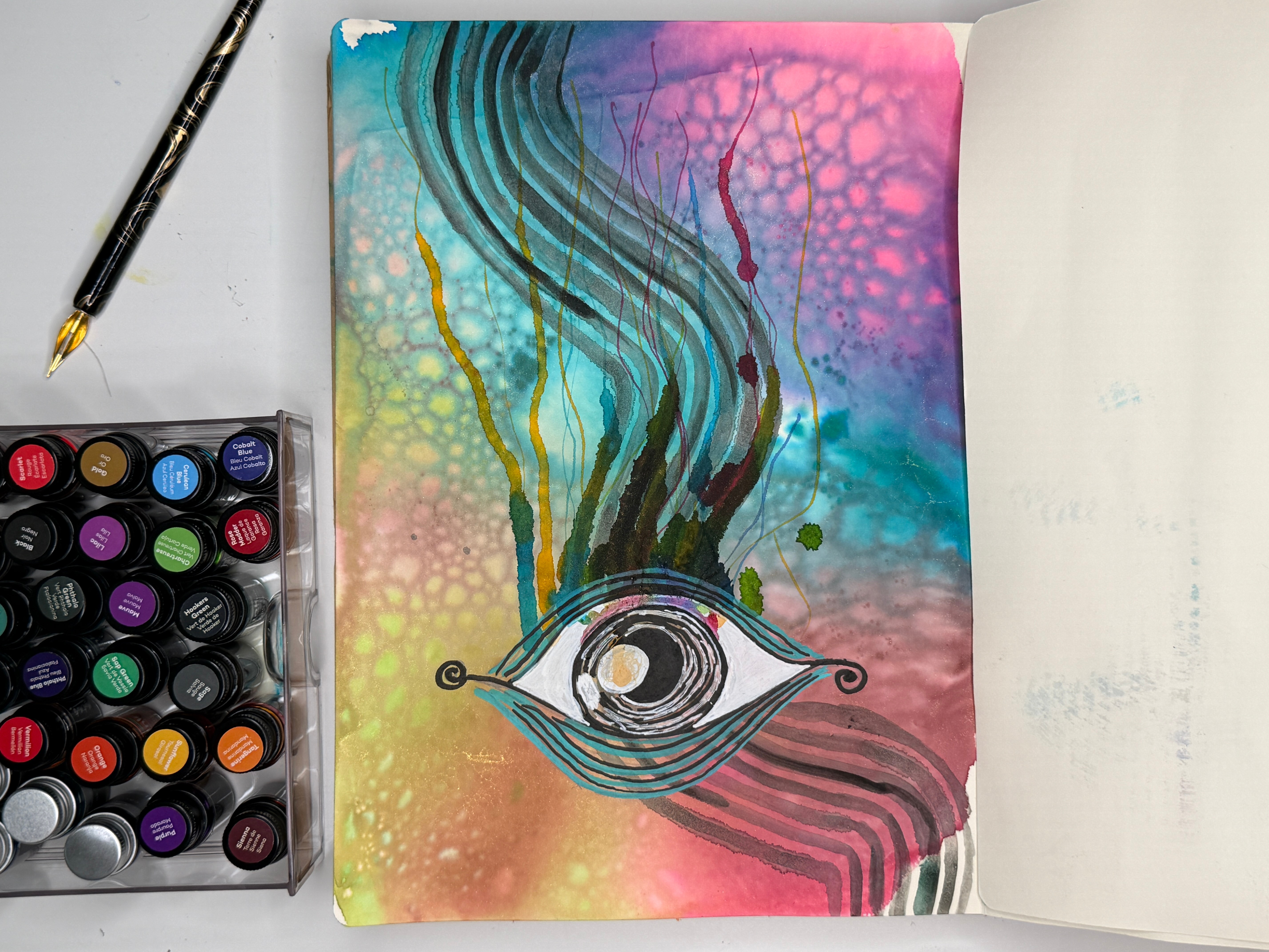 Colorful abstract painting with swirling lines and a central eye-like shape, on a notebook page, with paint bottles nearby.