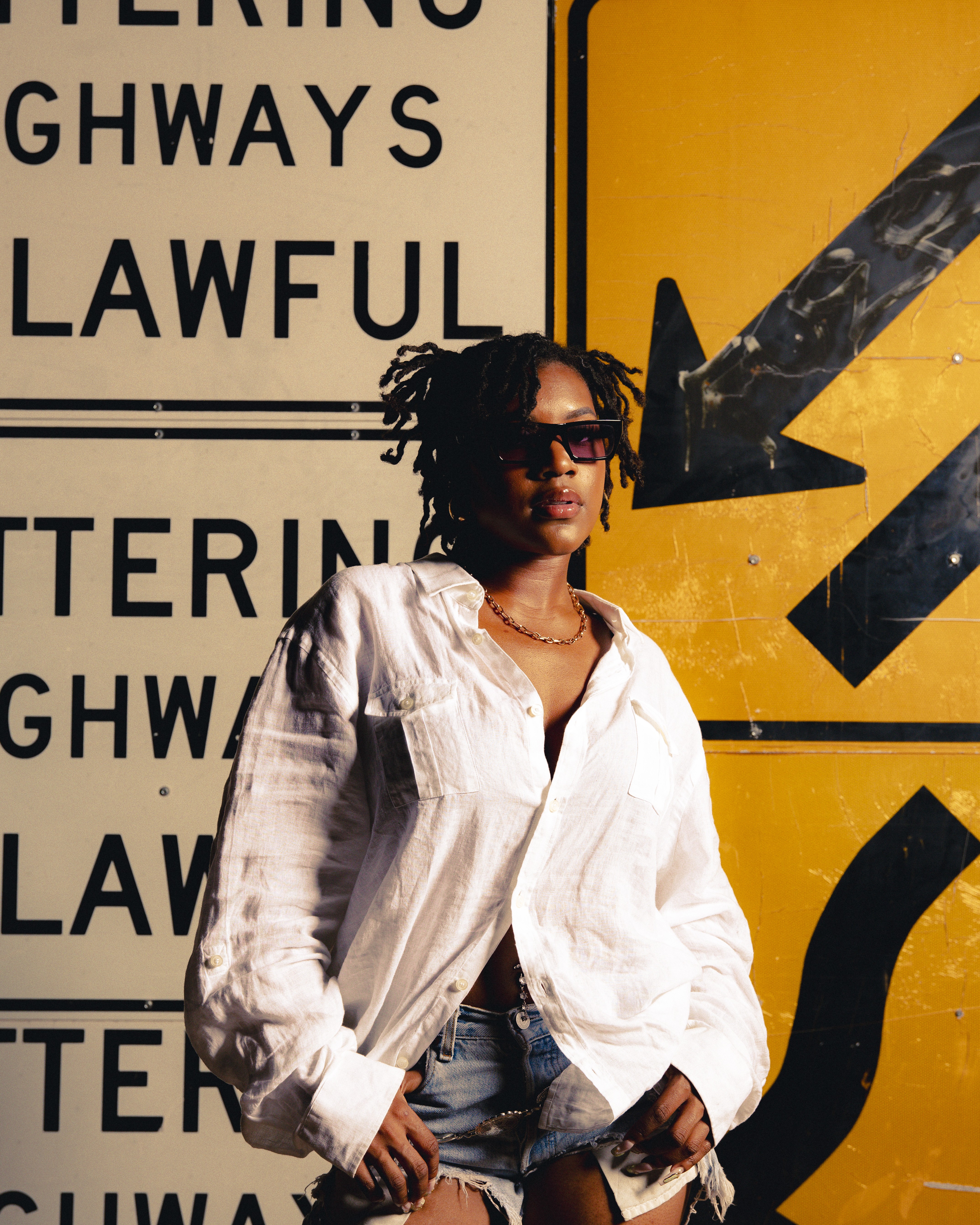 Person with sunglasses and curly hair standing in front of yellow and black road signs, wearing a white shirt and jeans.