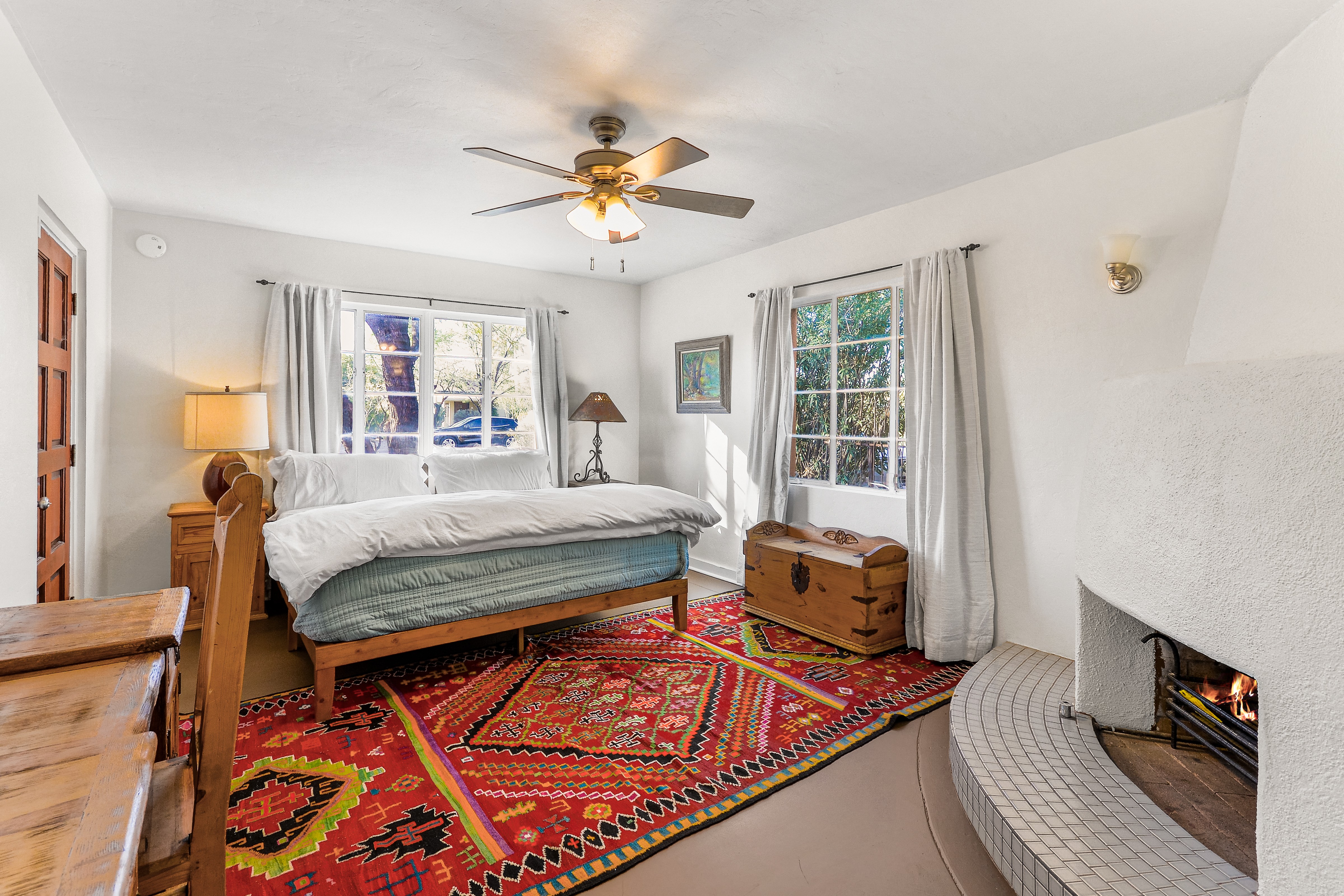 Bedroom with a bed, windows, a chest, and a fireplace, decorated with a colorful rug and curtains.