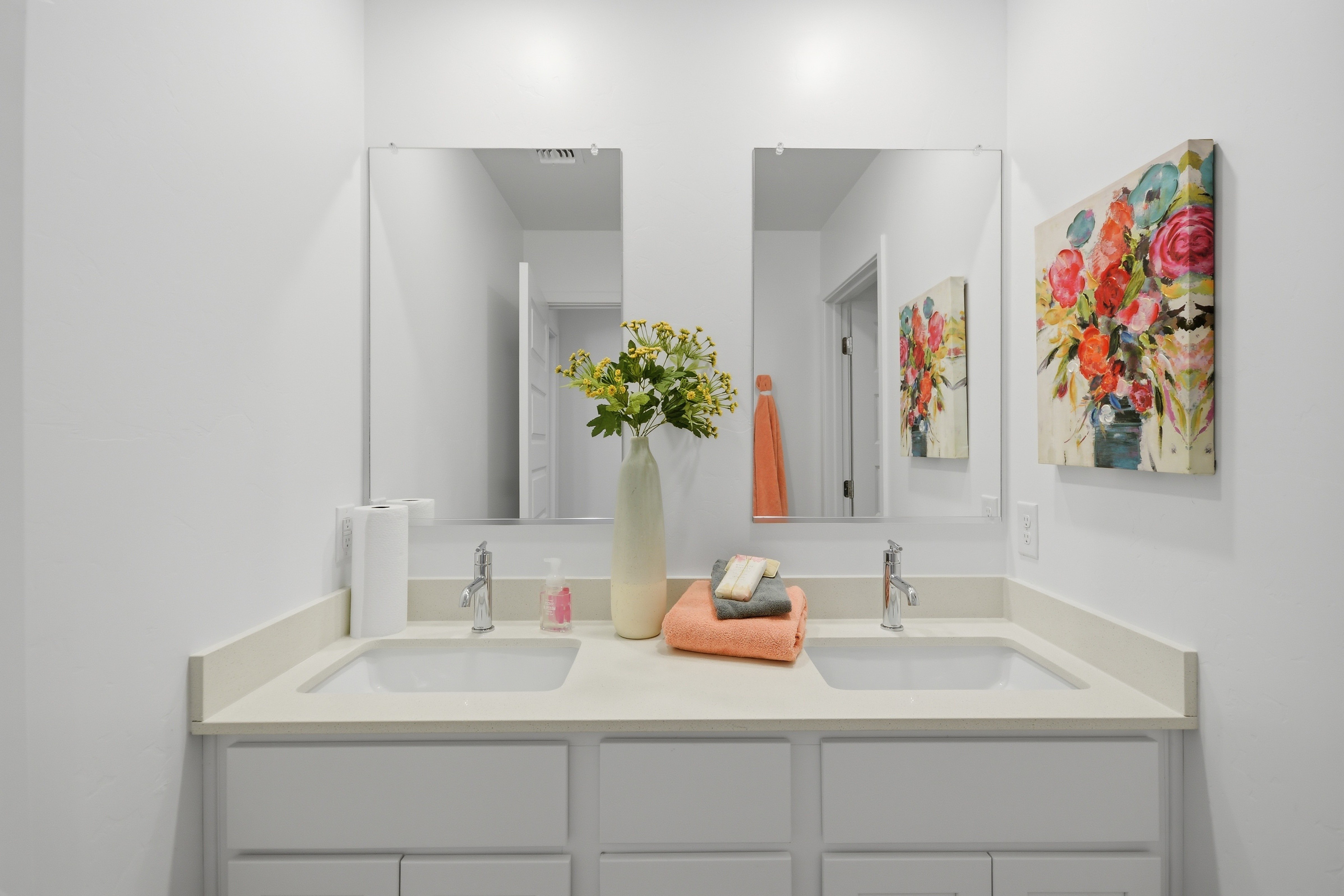 Bathroom vanity with two sinks, large mirror, flower vase, towels, and artwork on white walls.
