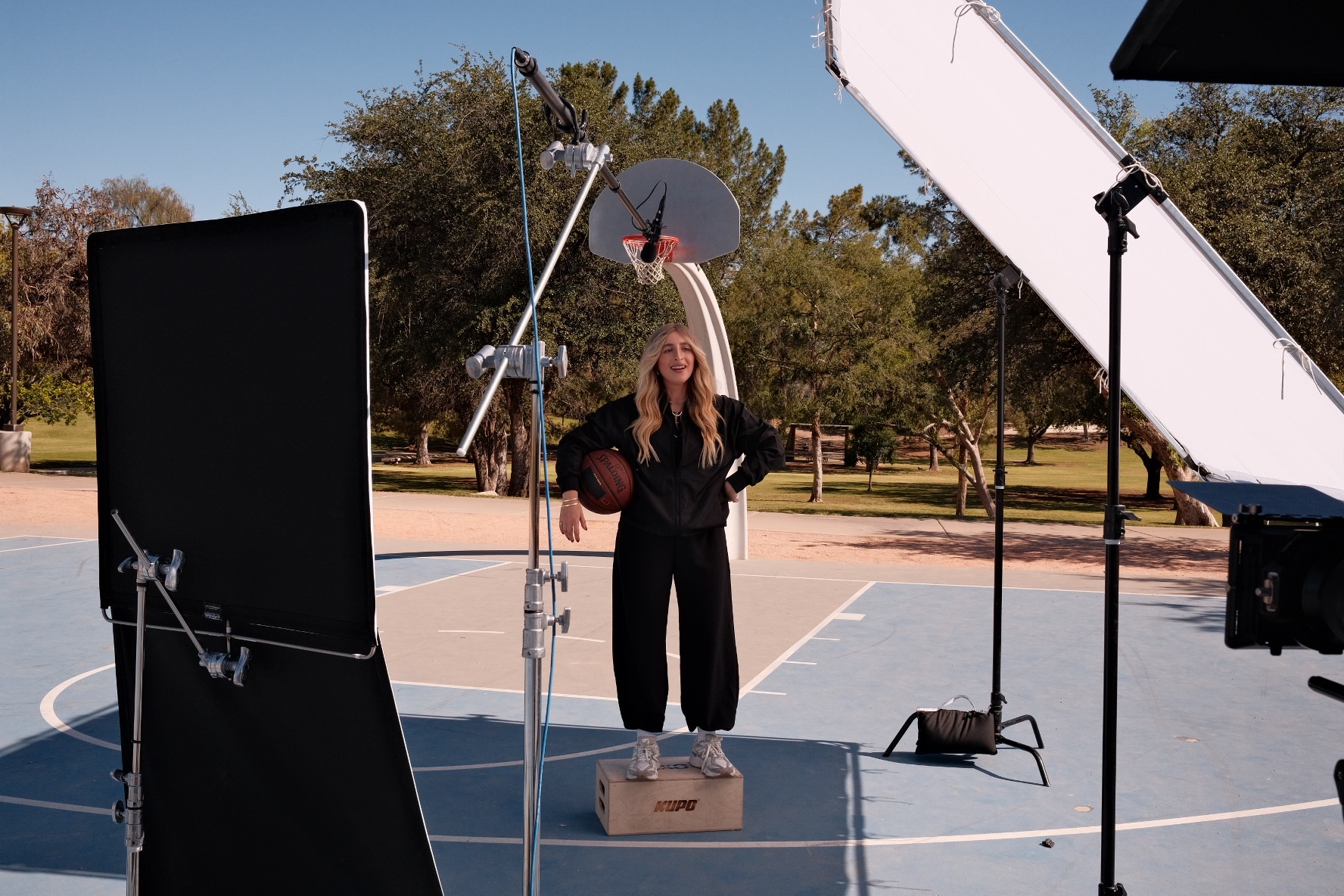 Person standing on a box in an outdoor basketball court, surrounded by photography equipment and lighting, with trees in background.
