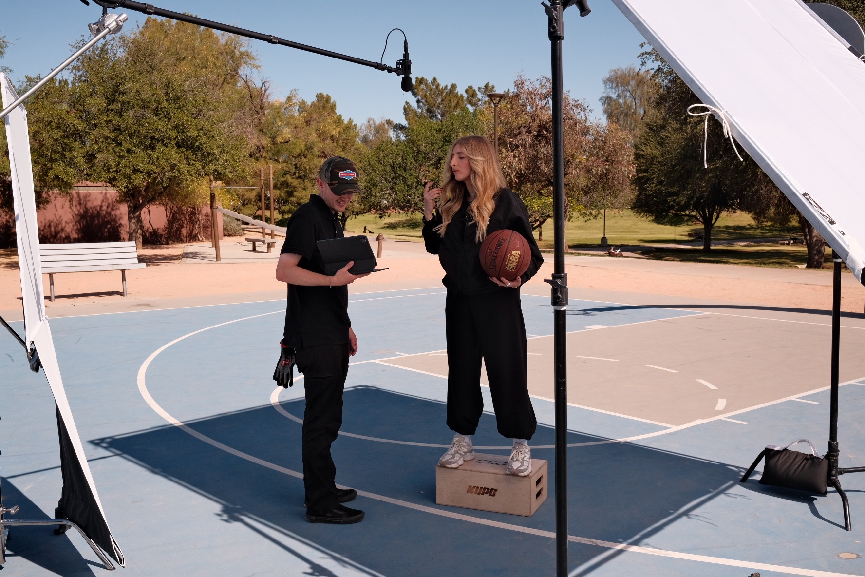 Two women on a basketball court, one holding a basketball, surrounded by lighting equipment, outdoors with trees and blue sky