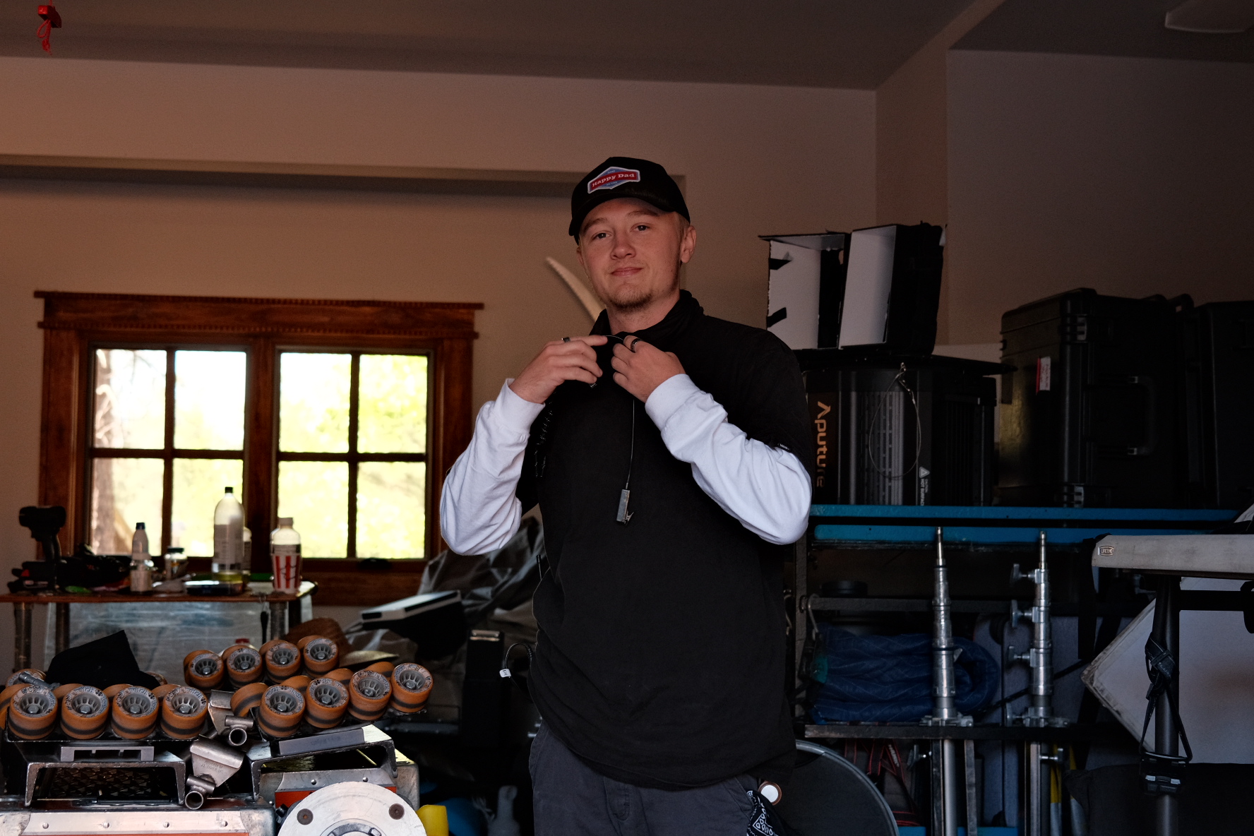 Young man wearing a cap and black jacket standing indoors with a window behind him, holding a small object near his face.