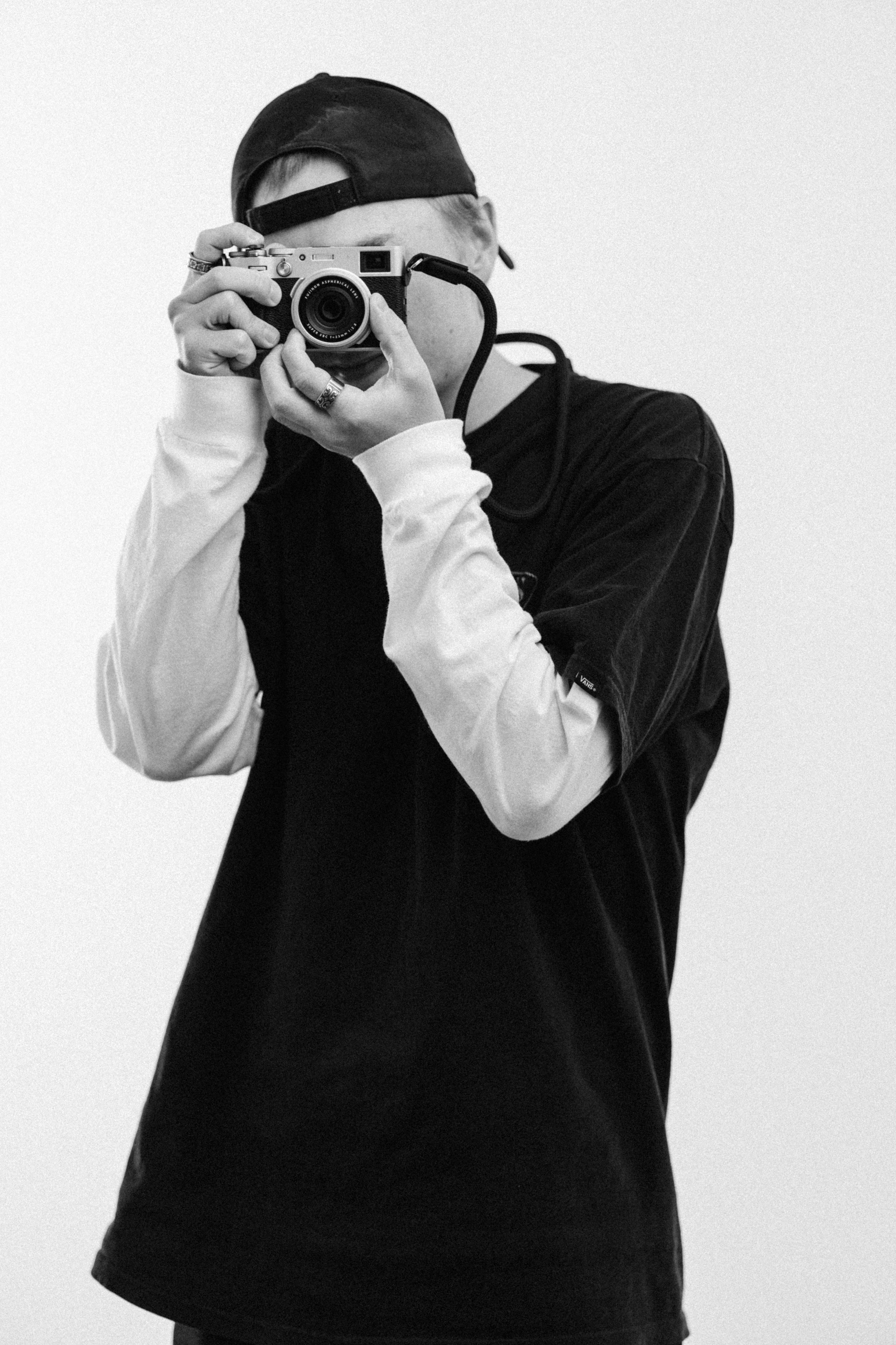 Person wearing a cap and black and white clothing takes a photo with a camera, facing forward.