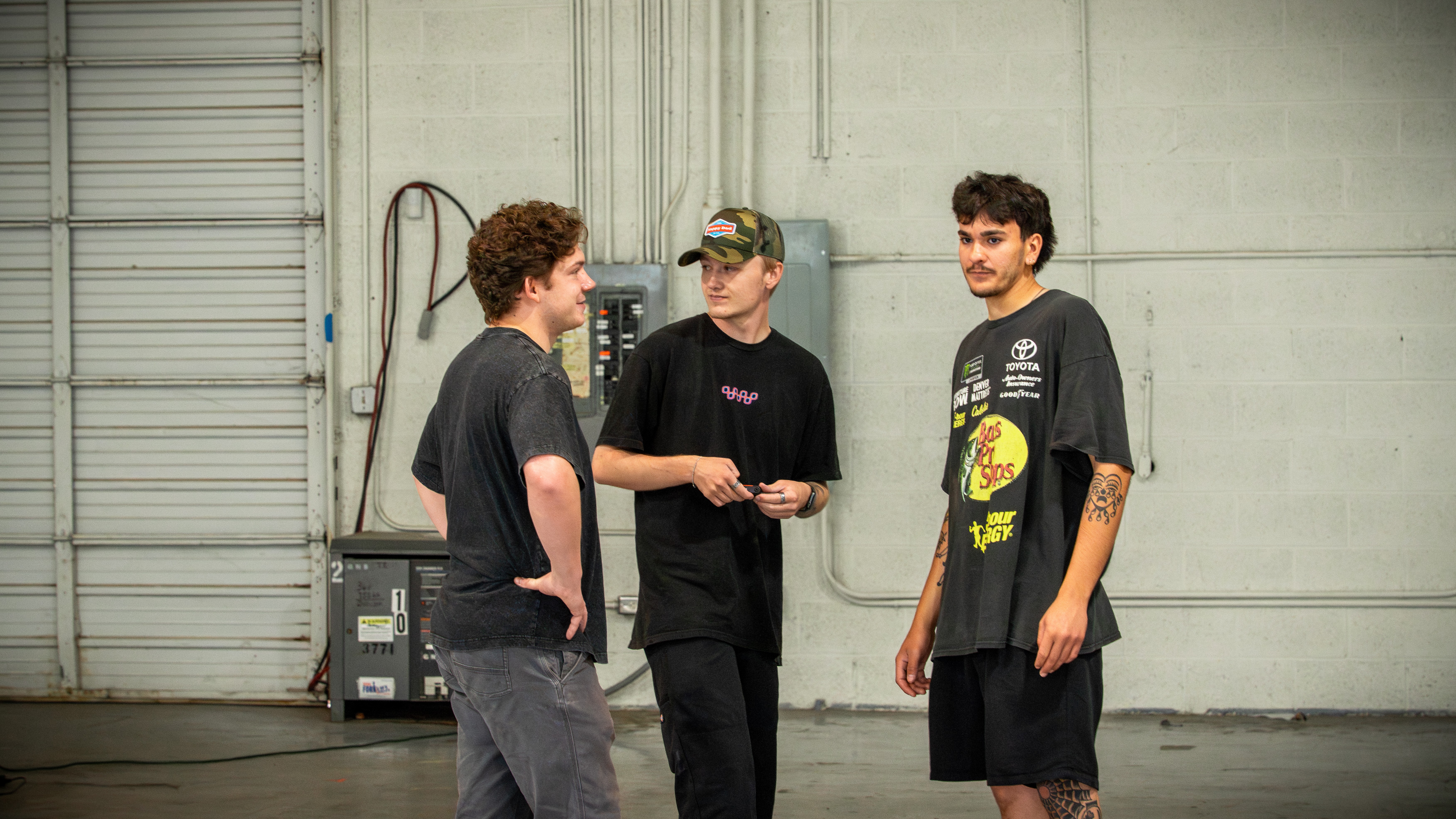 Three young men standing and talking in an industrial space with a large garage door in the background.