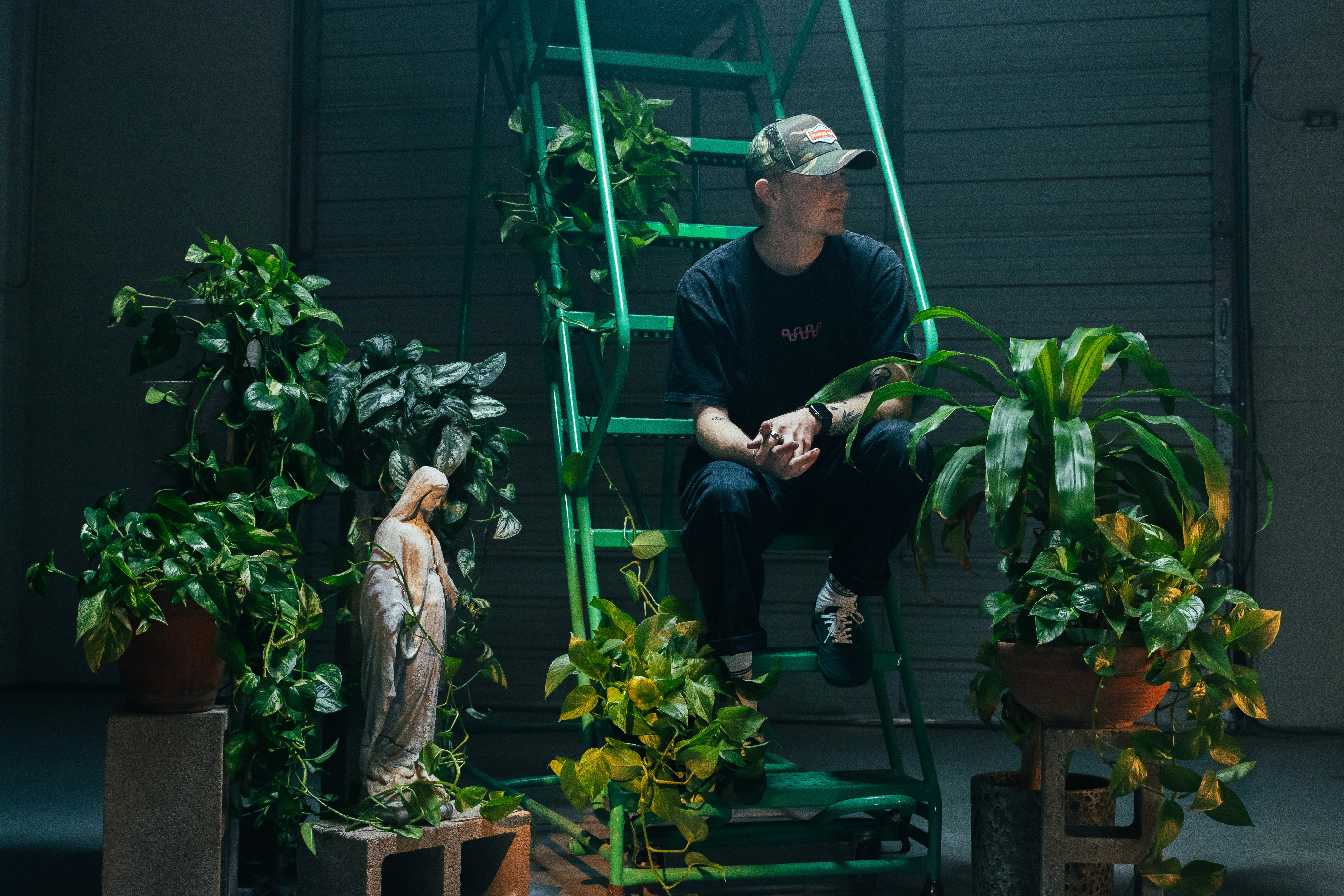 Person sitting on a ladder surrounded by potted plants and a statue, in a garden or indoor space.