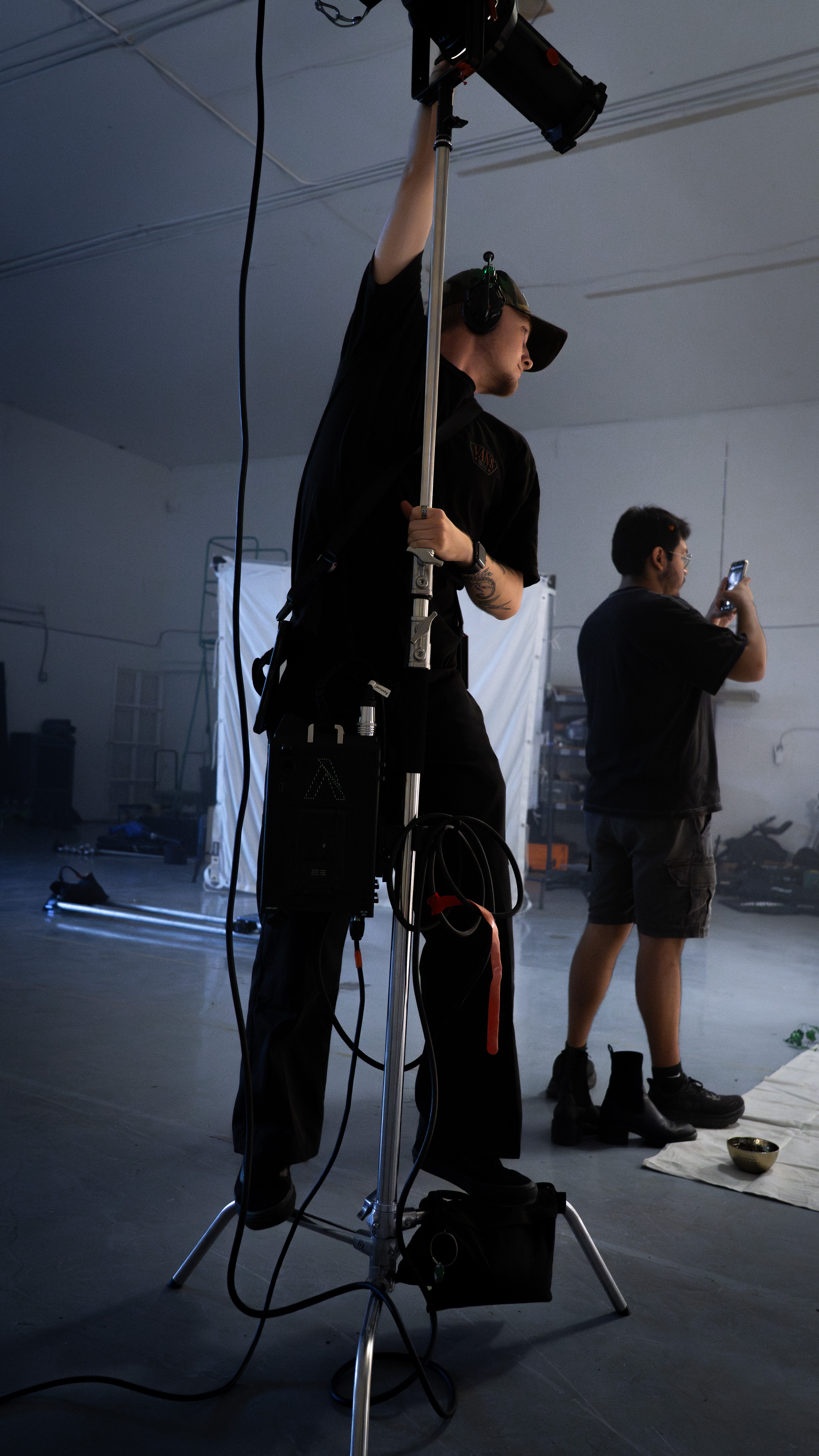 Person adjusting equipment in a studio with another person taking a photo in the background.