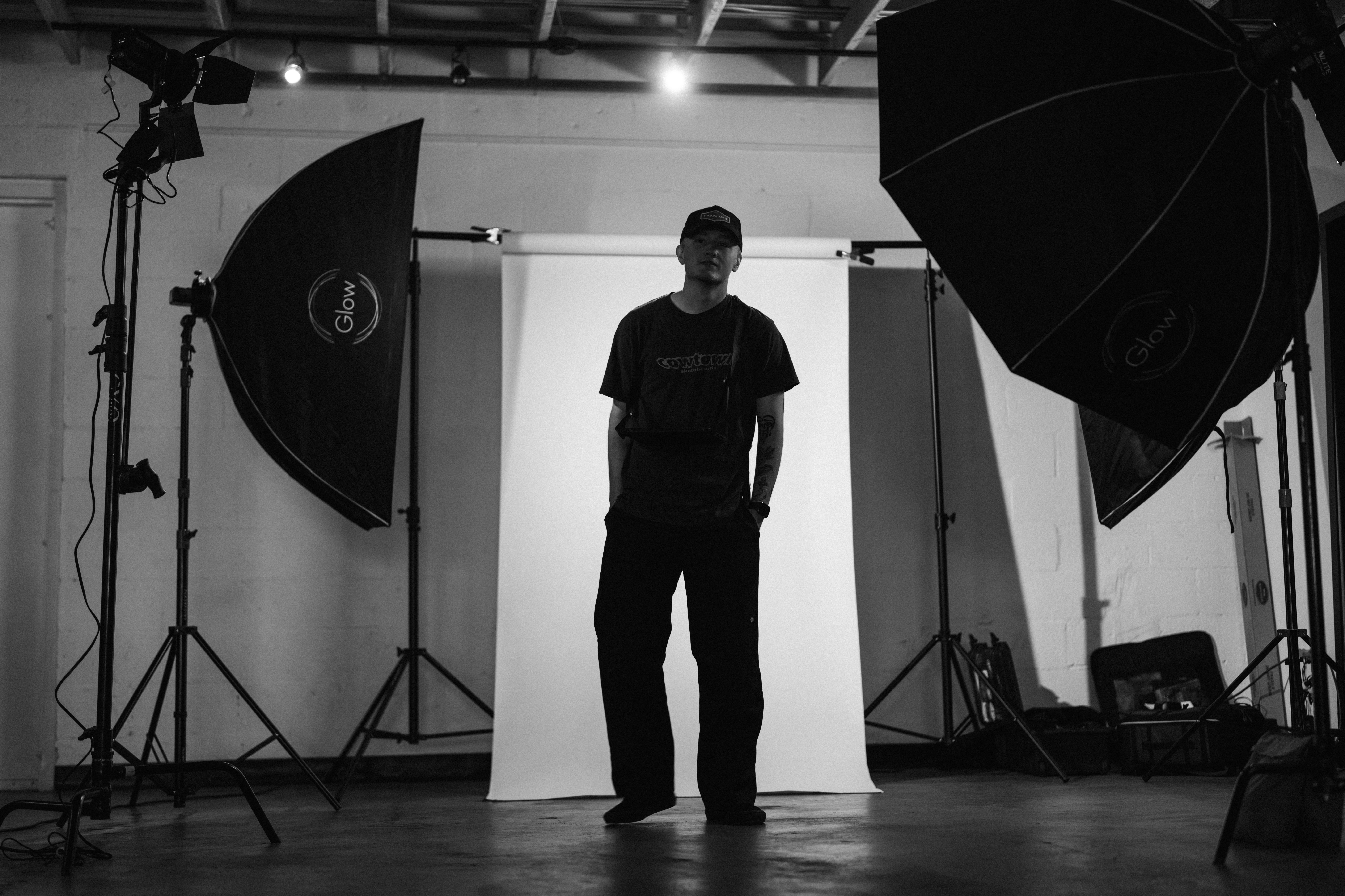 Person standing in a photography studio with lighting equipment and backdrop, facing forward, in black and white.