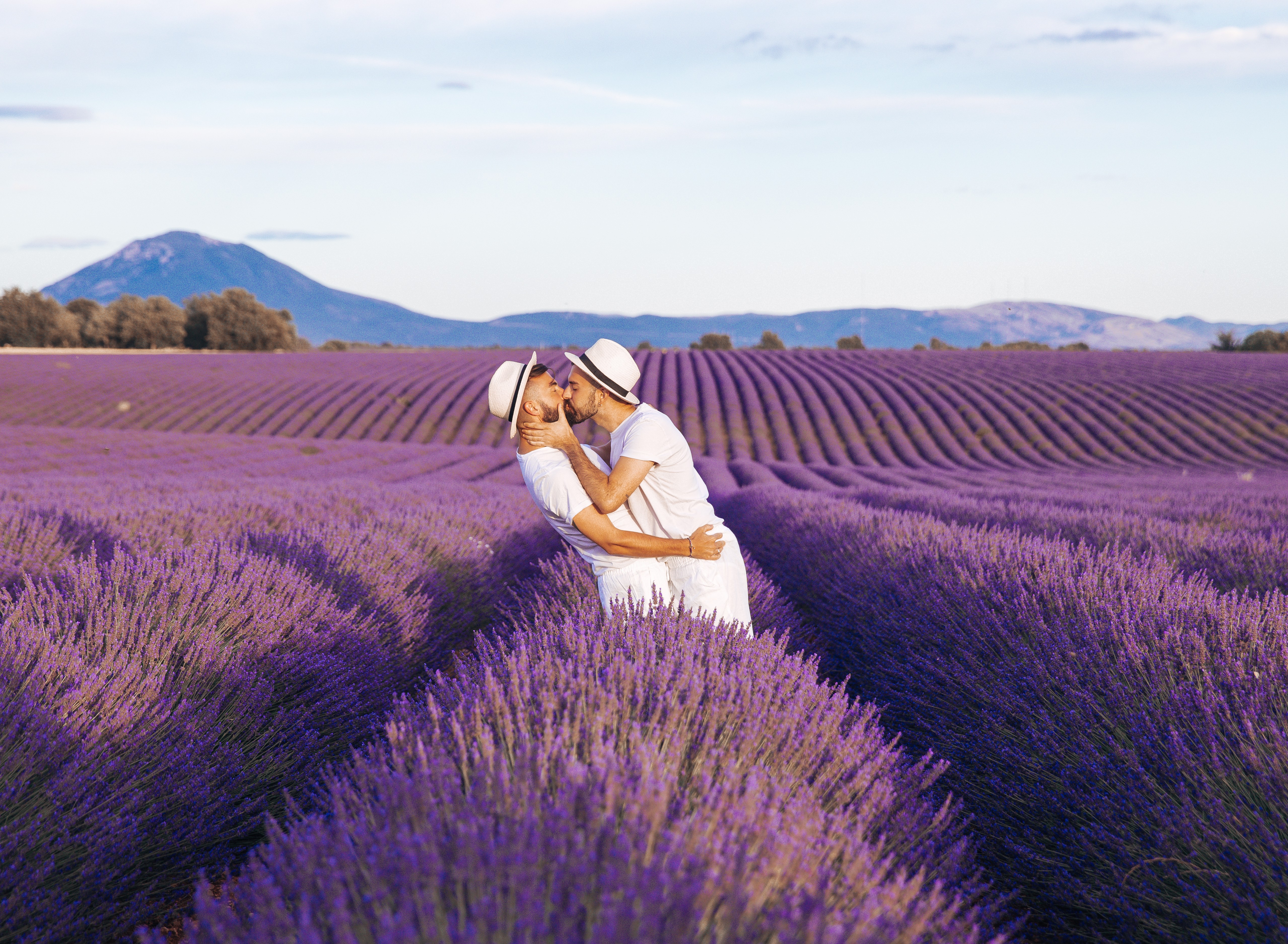 A couple embraces in a lavender field with mountains in the background, both wearing white and hats.