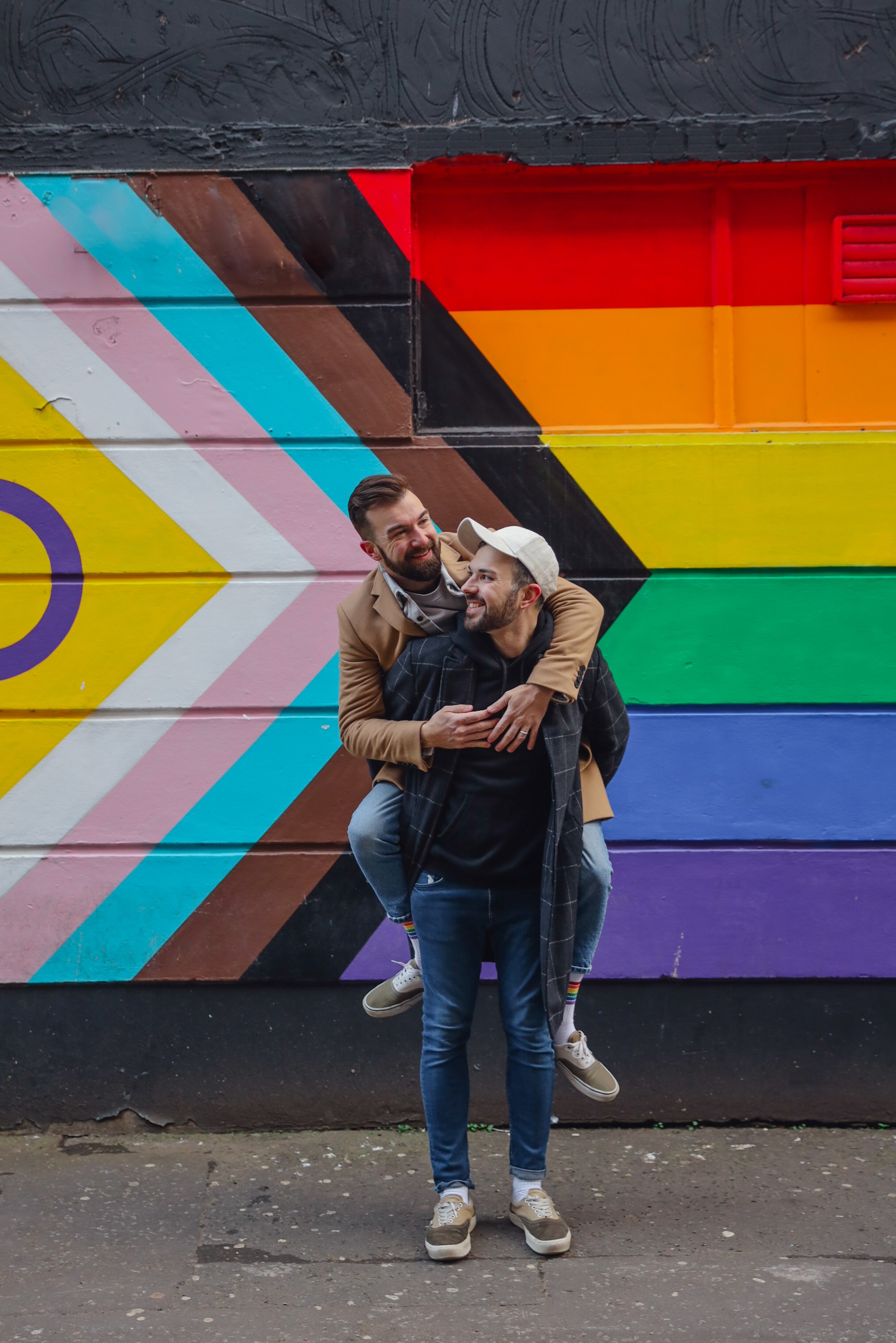 Two people hugging in front of a colorful rainbow mural wall, one wearing a cap and the other with glasses, smiling.