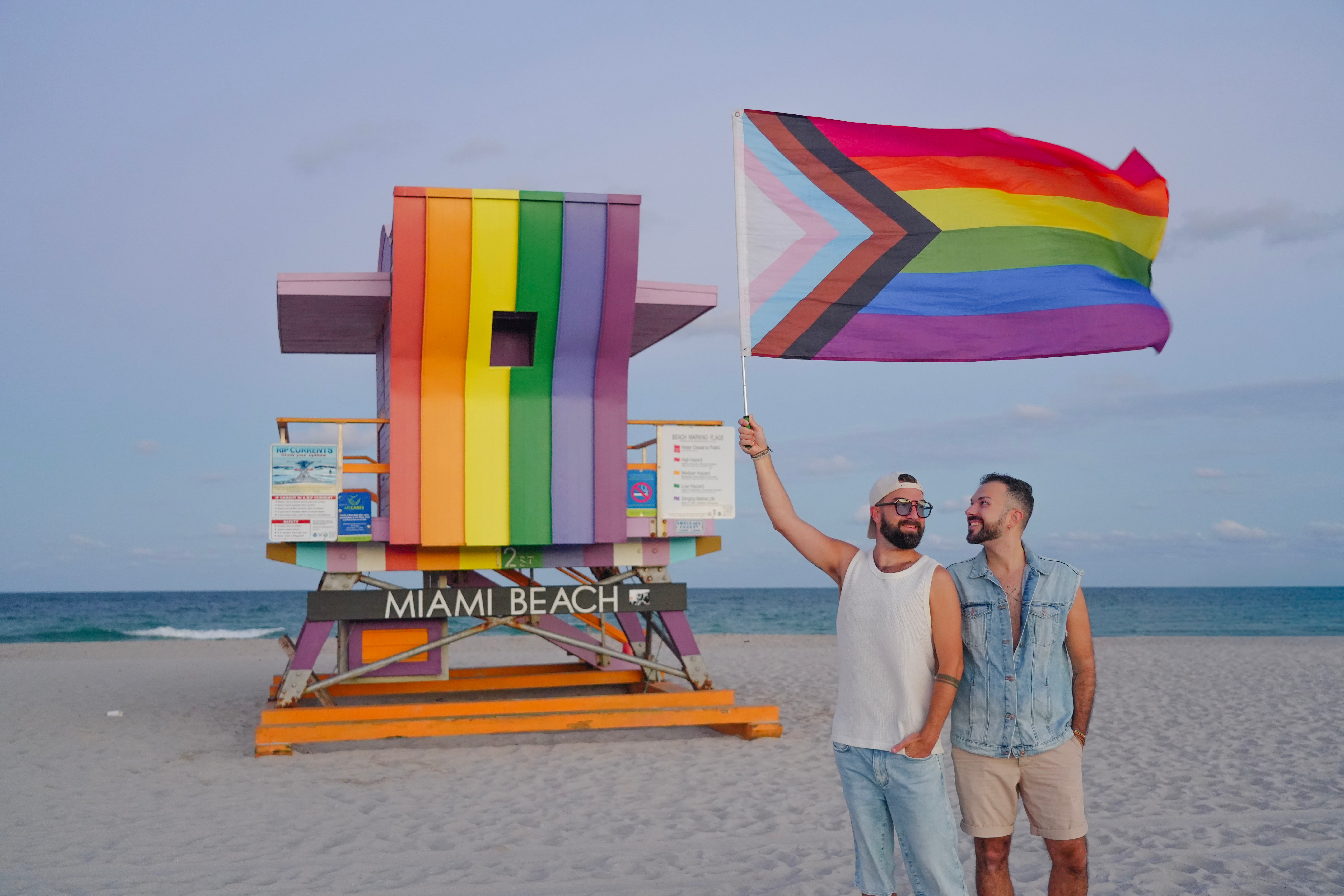 Two men stand on the beach near a rainbow flag and a lifeguard tower with rainbow decorations, holding a rainbow flag.