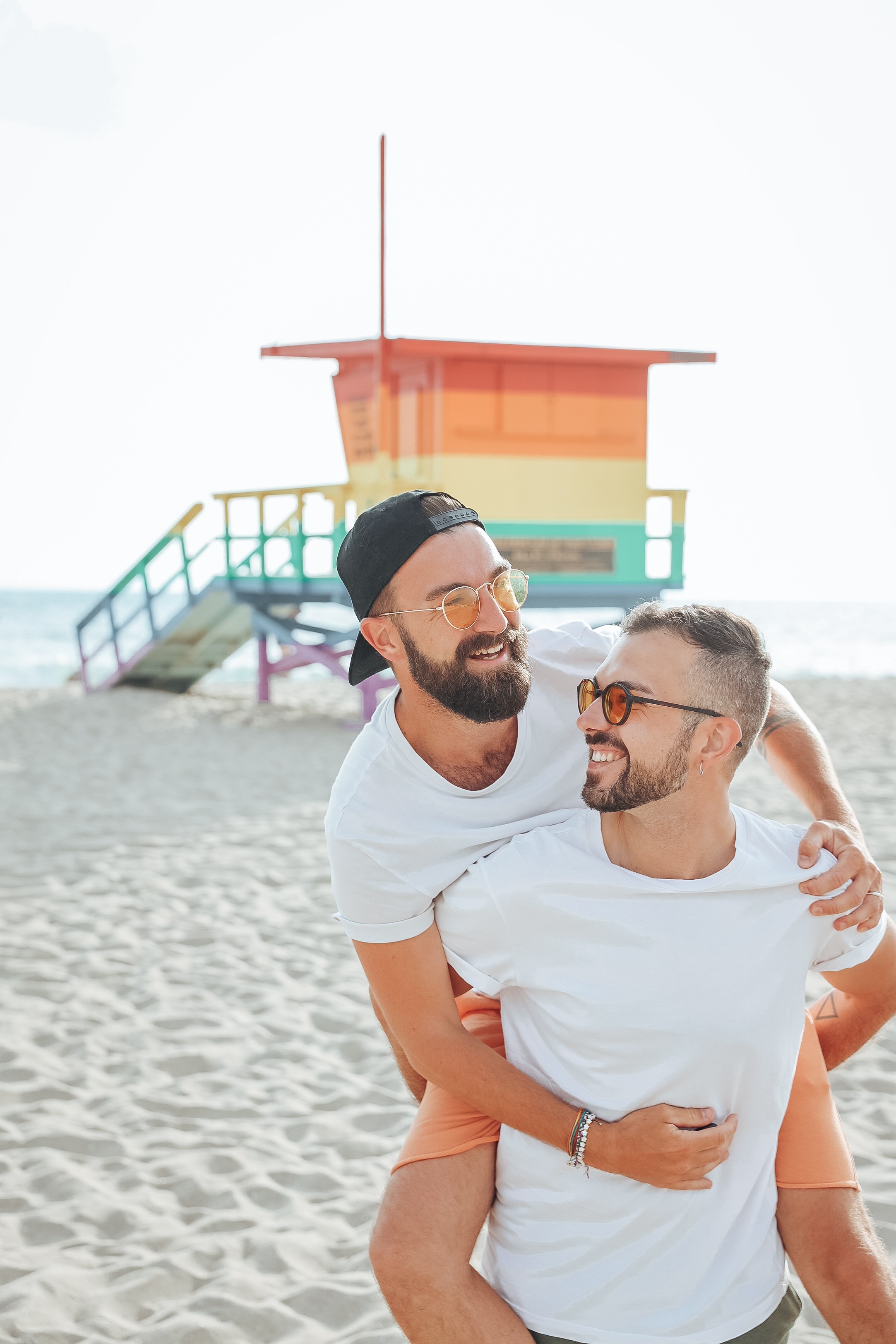 Two men smiling and hugging on the beach with a colorful lifeguard tower in the background.